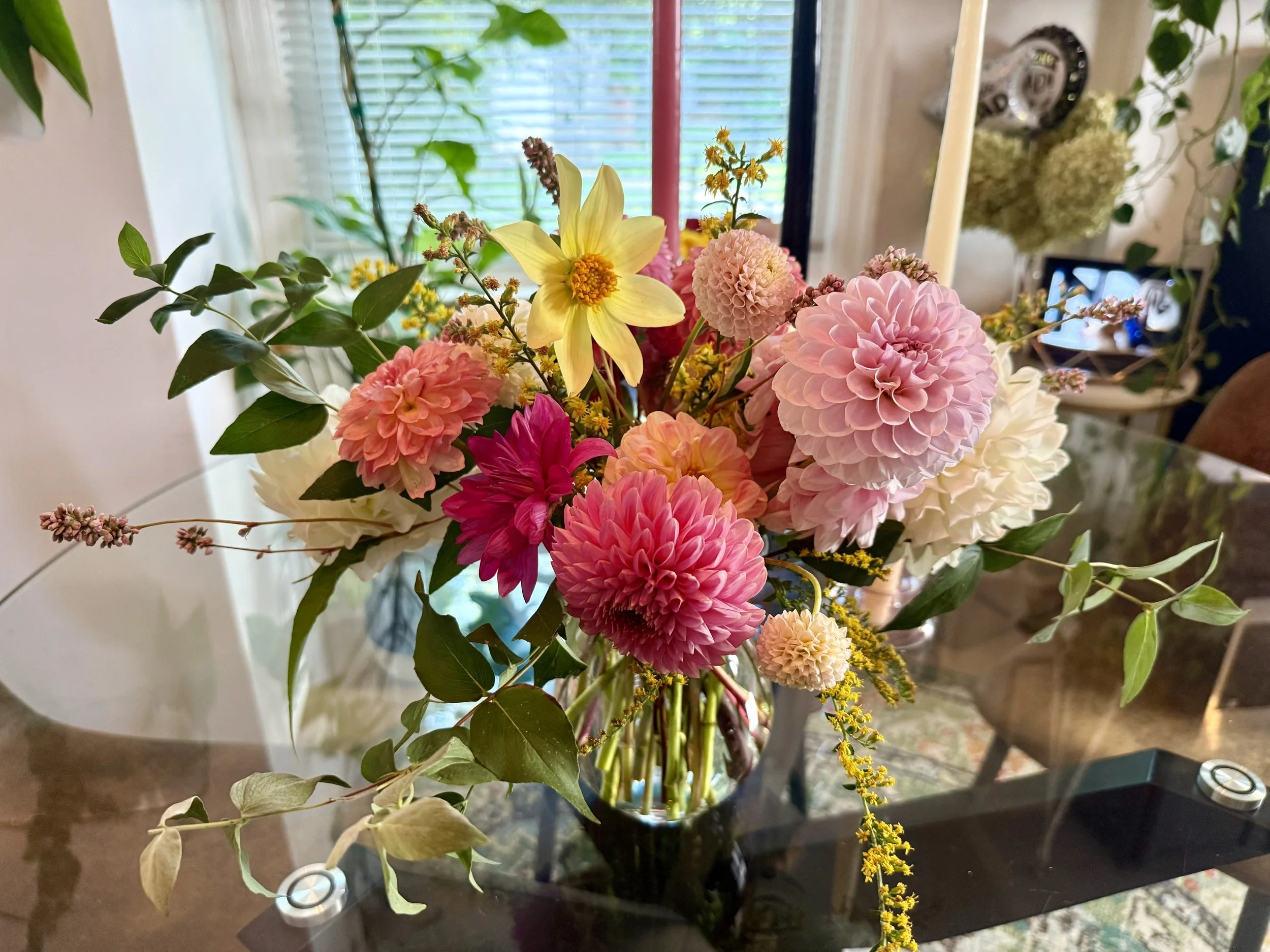 Colorful bouquet of pink, yellow, and white flowers with green leaves arranged in a glass vase on a glass table in a bright room.