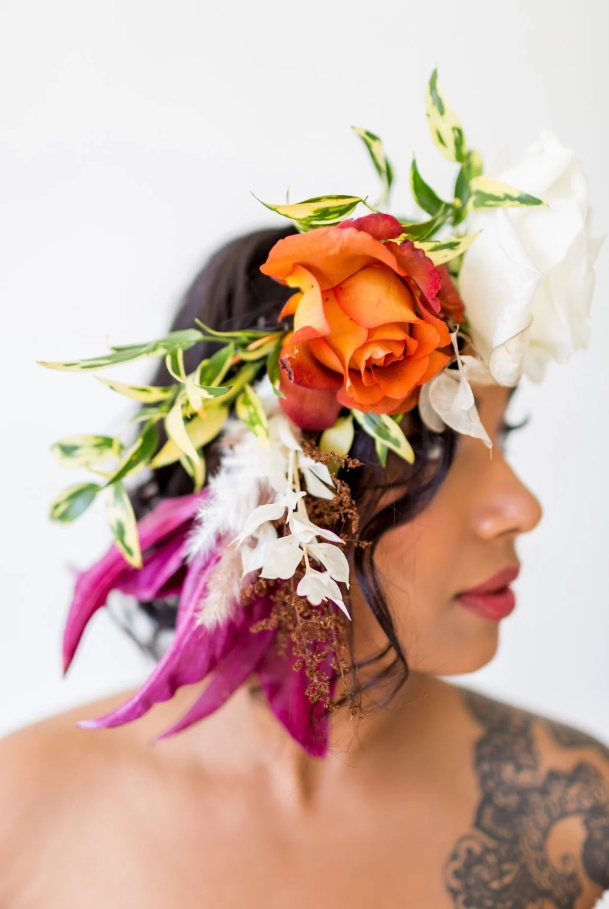 Side profile of a woman with dark hair and colorful floral headpiece, featuring orange, white, pink, and green flowers and leaves, against a plain light background.