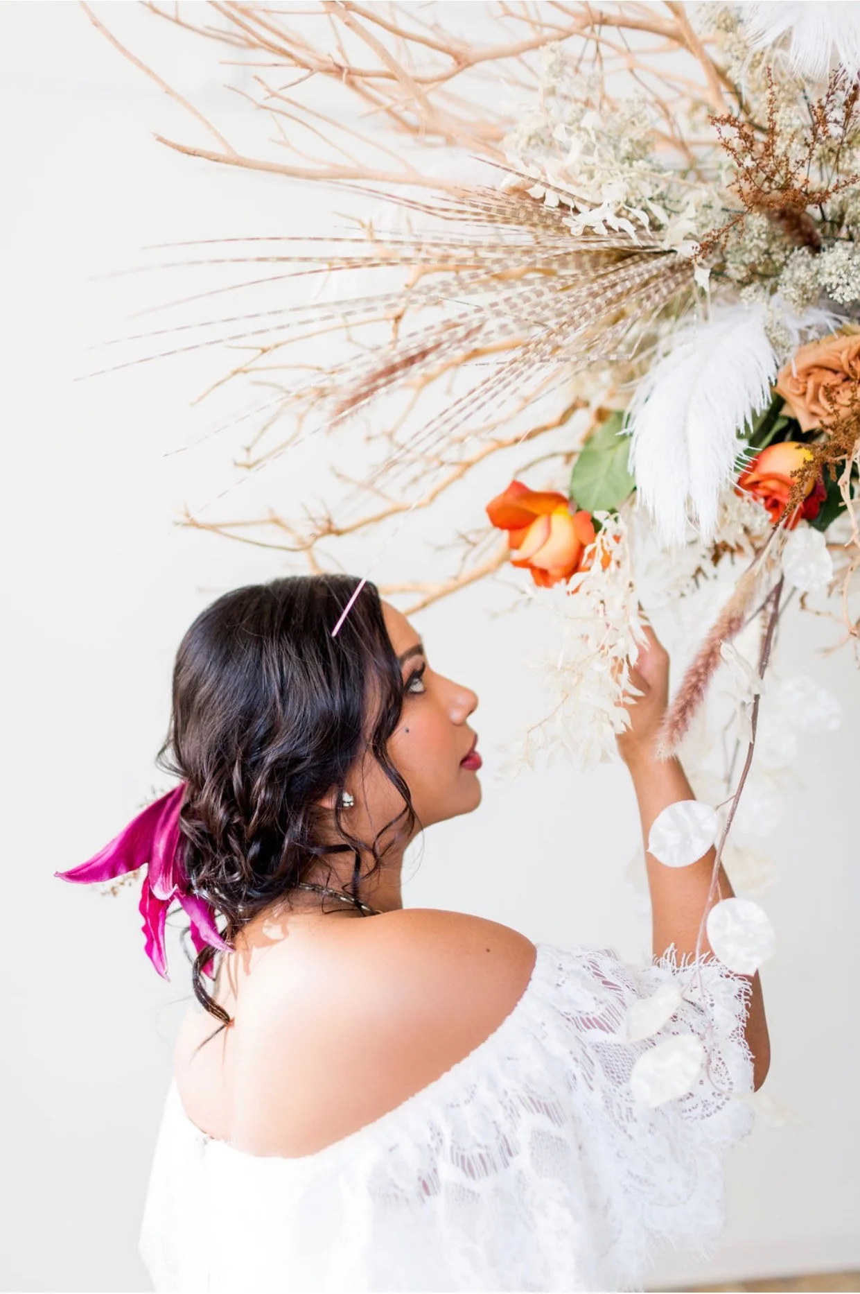 A woman with dark wavy hair and a pink ribbon in her hair decorates a large floral arrangement with orange roses, white feathers, pampas grass, and dried branches.