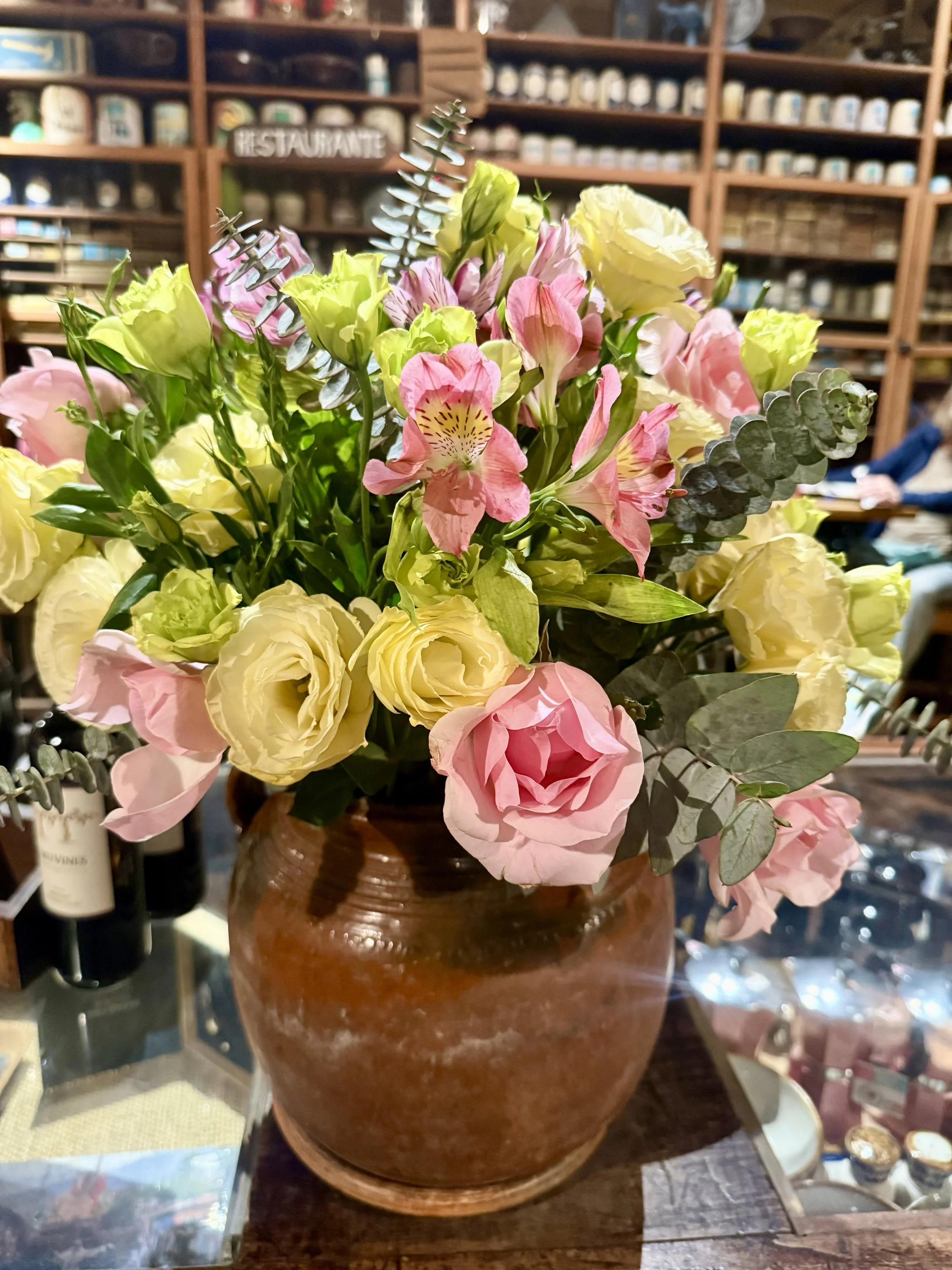 A ceramic vase filled with pink, cream, and green flowers, including roses, alstroemeria, and greenery, placed on a wooden surface in a store with shelves of jars and bottles in the background.