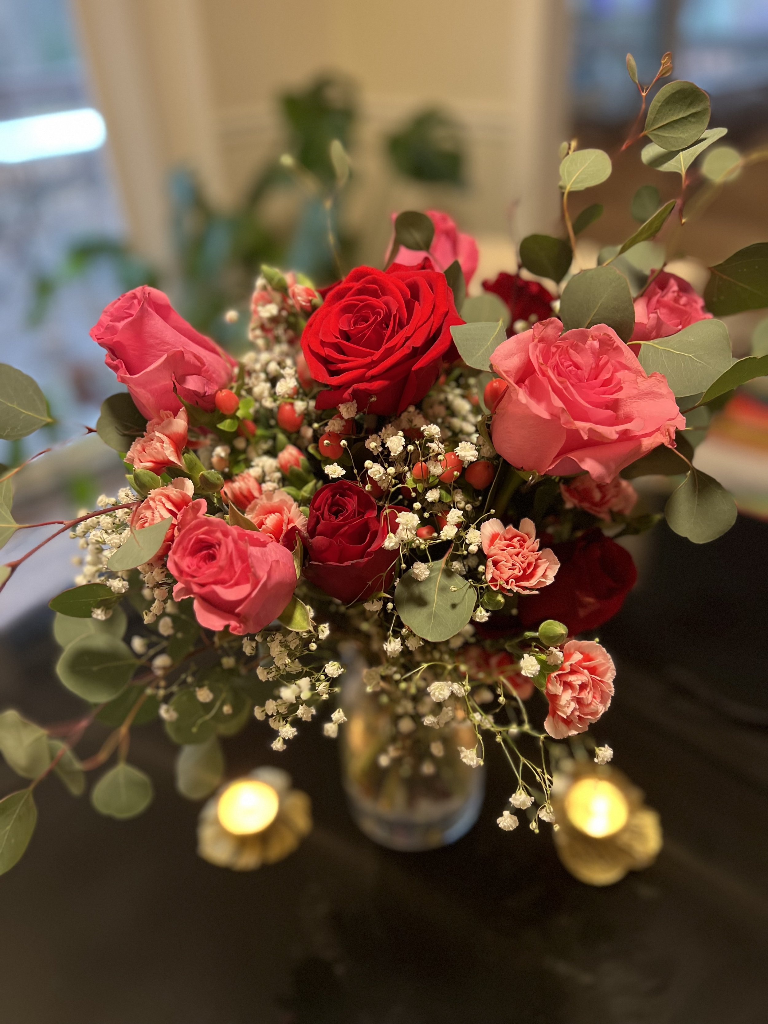 A vase of mixed pink and red roses, baby's breath, and greenery, placed on a dark surface with two small lit candles nearby.