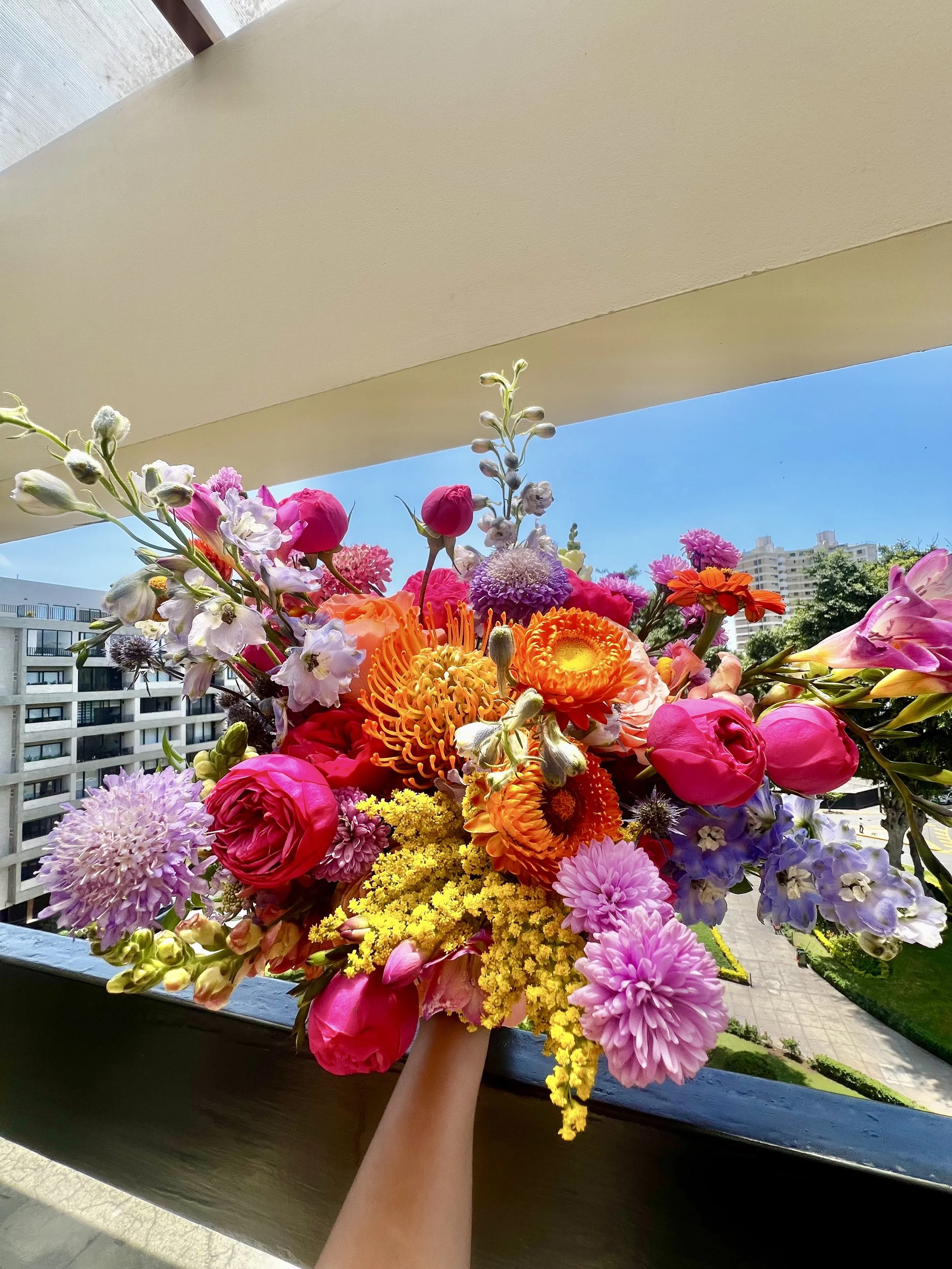 Colorful bouquet of various flowers being held outdoors, with buildings and a clear blue sky in the background.