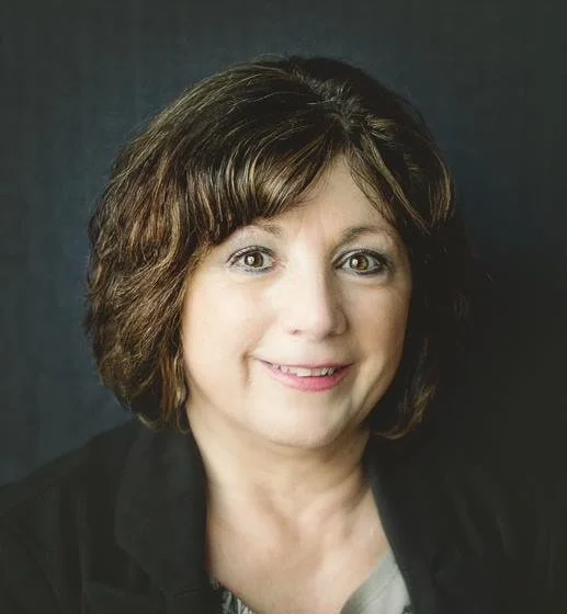Helen Wean Headshot. Close-up of a woman with short, wavy brown hair and light makeup, smiling against a dark background.