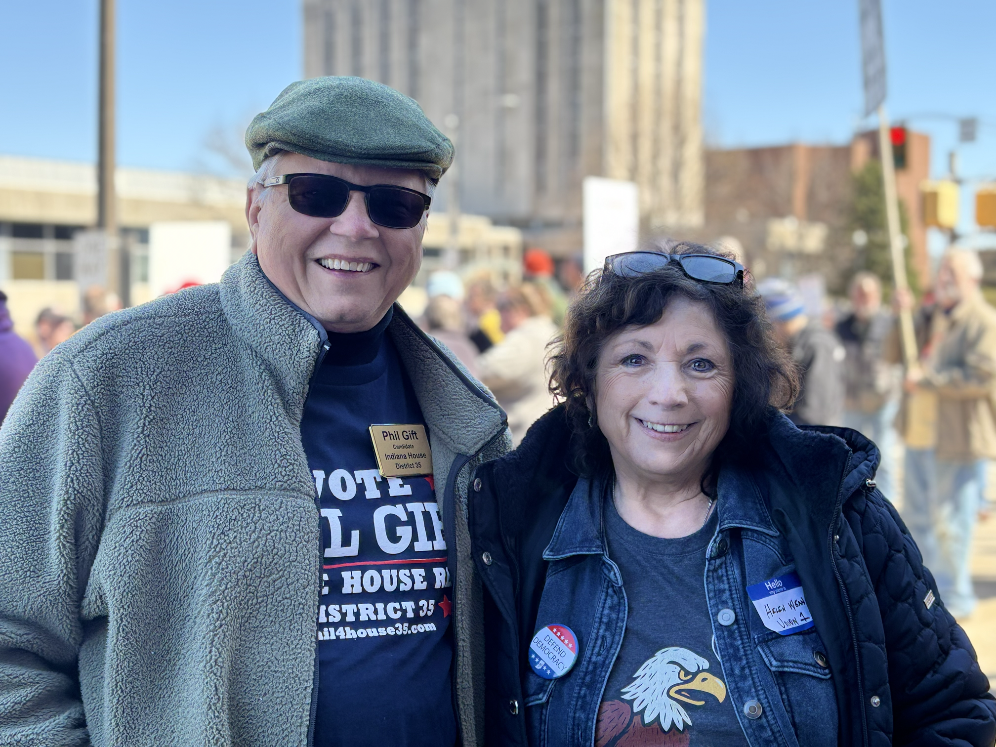Two smiling people, a man and a woman, standing outdoors at a rally. The man is wearing sunglasses, a flat cap, a fleece jacket, and a campaign button with the text "Phil Gitt Candidate Indiana House District 35." The woman has curly hair, glasses on her head, and is wearing a denim jacket, a T-shirt with a bald eagle graphic, and a campaign button. The background shows a crowd of people, some holding signs, and city buildings.