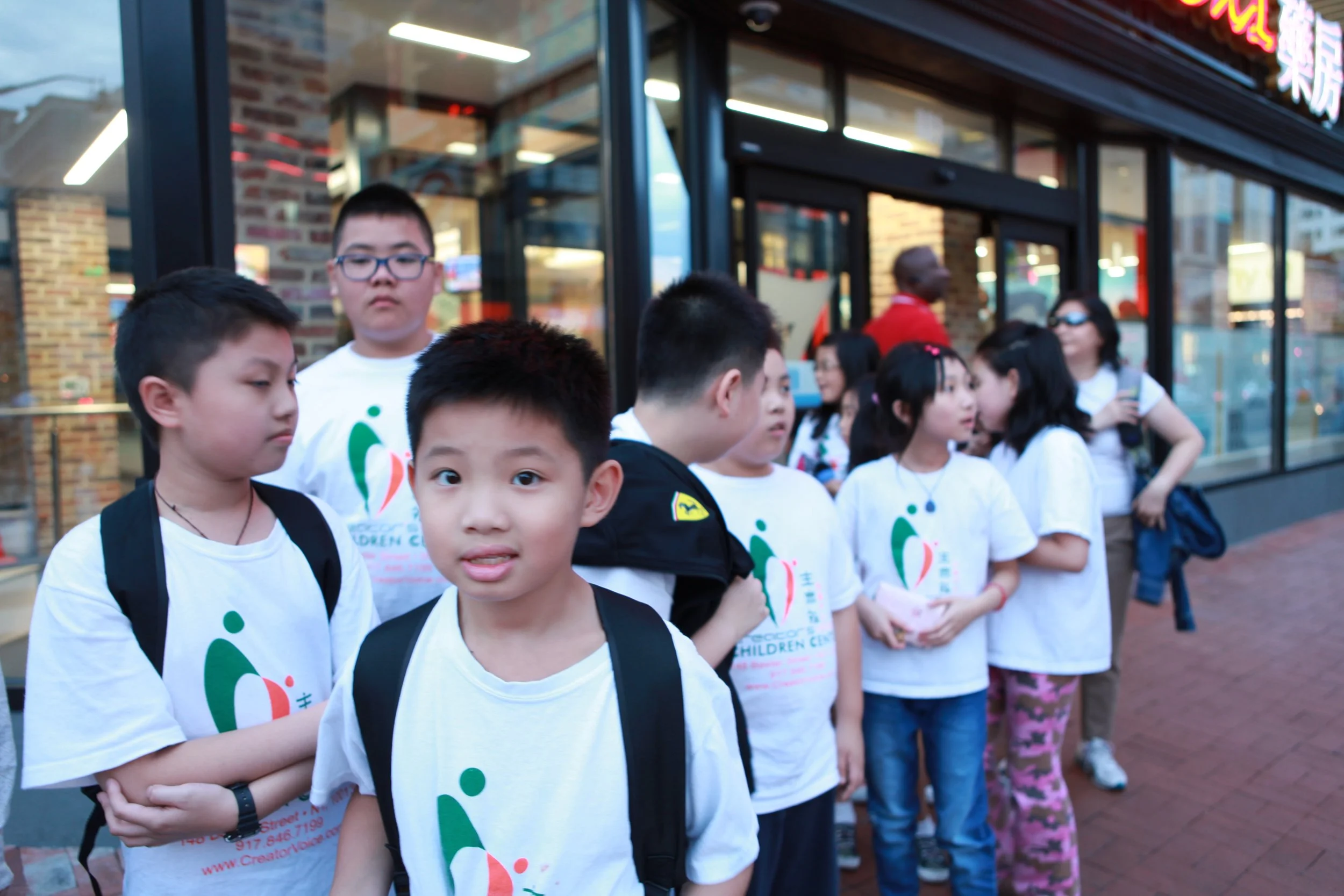 A group of children, mostly boys and girls, standing in line outside a building with glass windows and brick walls. They are wearing white t-shirts with colorful designs and text, possibly participating in a community or school event.