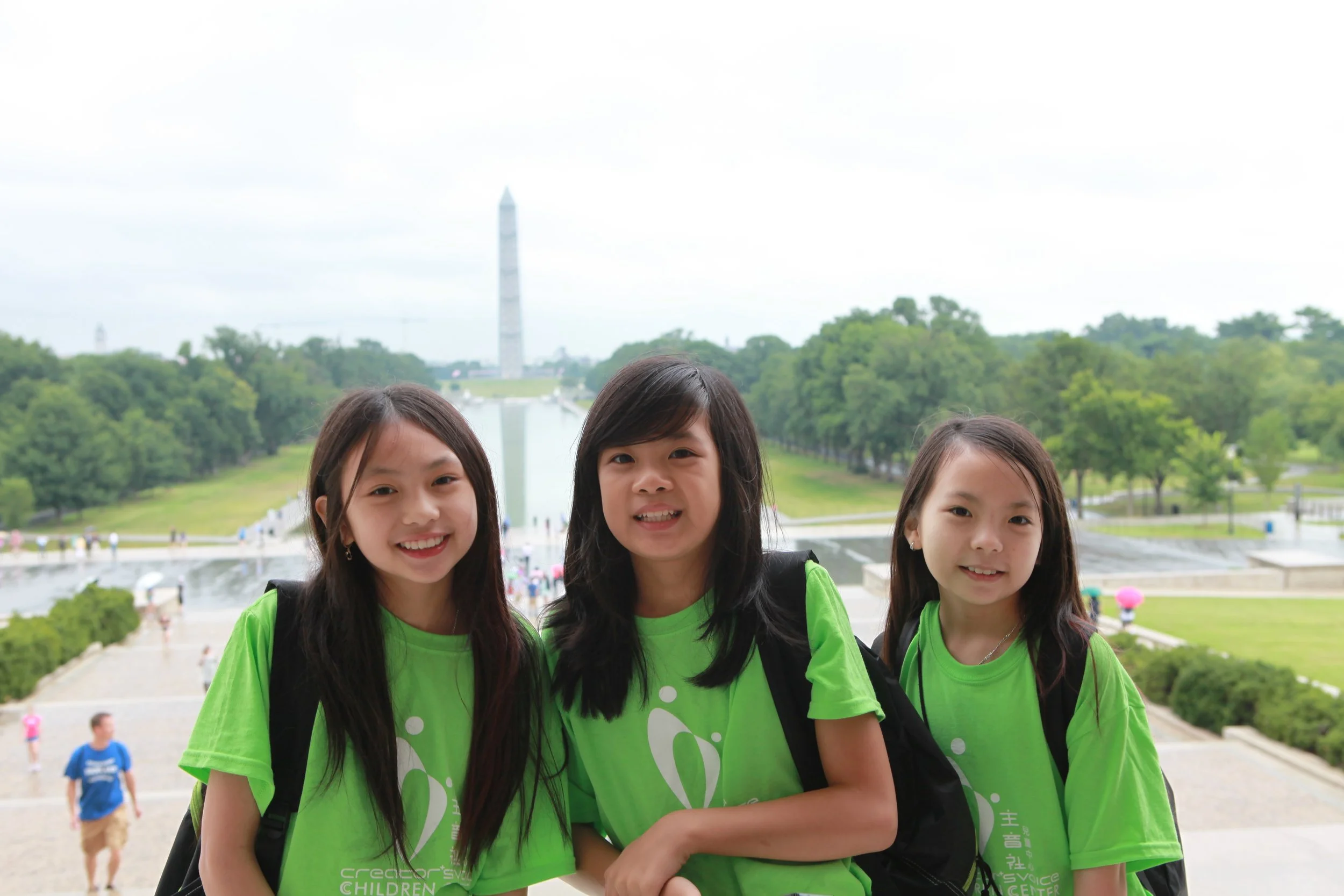 Three young Asian girls wearing bright green T-shirts with white text and graphics, standing outdoors in front of the Washington Monument and reflecting pool, with other visitors in the background.