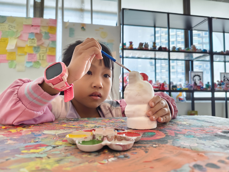 Young girl painting a ceramic sculpture in an art studio with shelves of colorful figurines in the background.