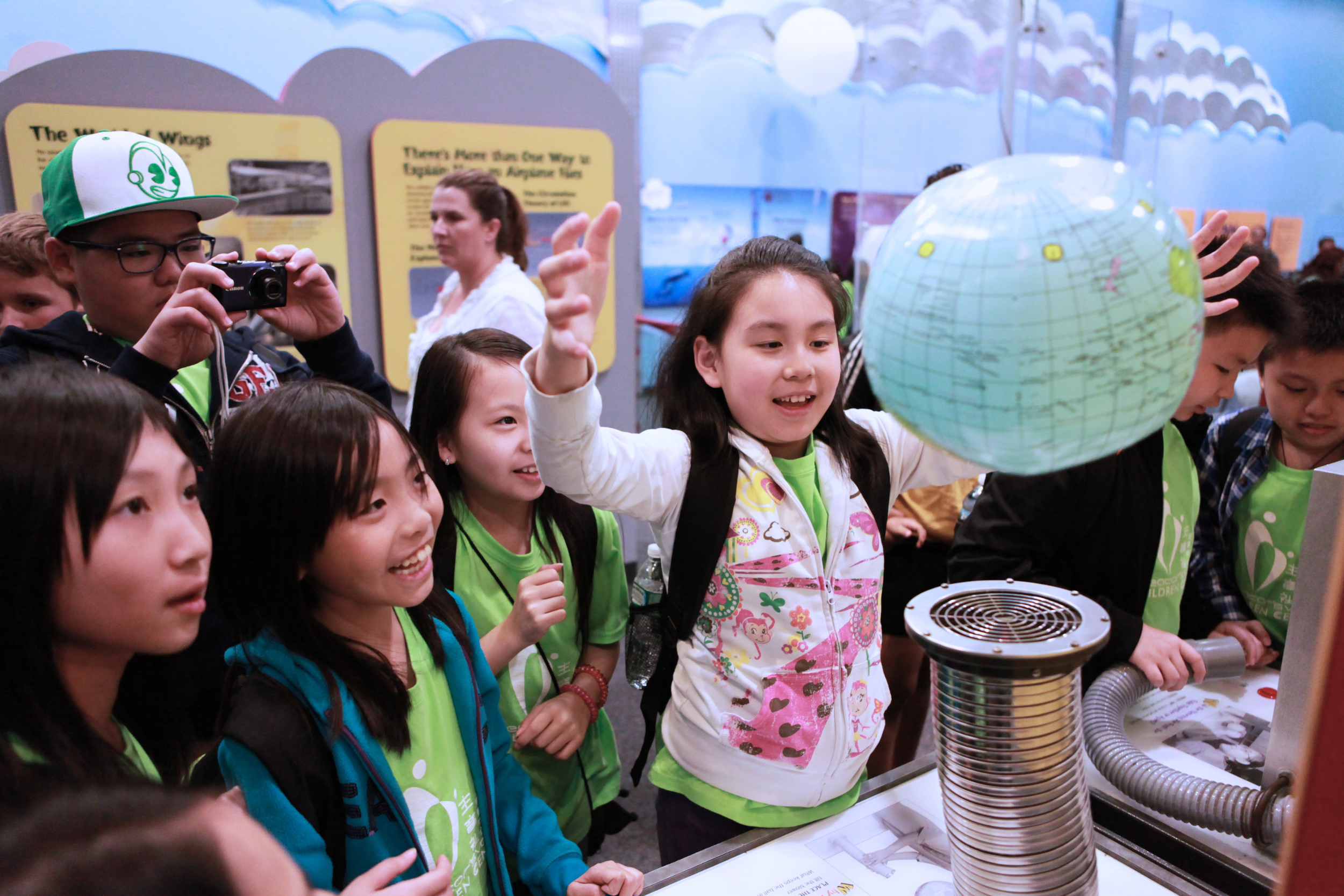 Children gathered around at a science exhibit, exploring a device with a globe and tubes, while an adult observes in the background.