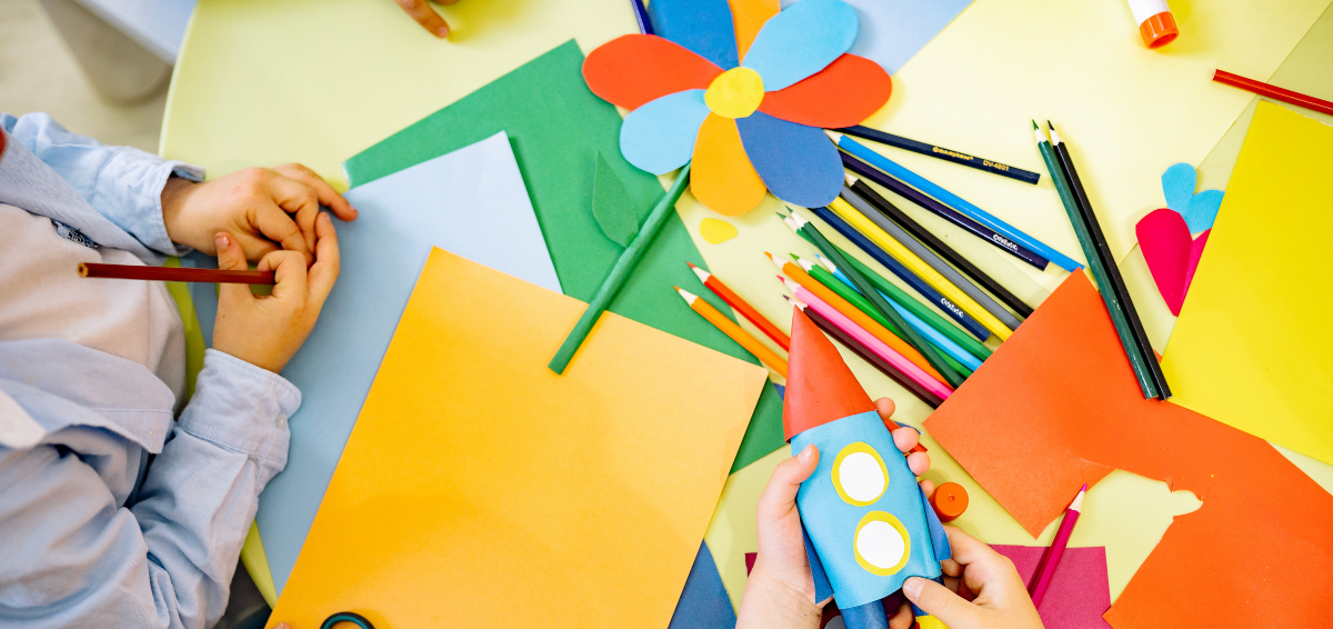 Child and adult working on paper craft project at a table with colored paper, markers, and a paper rocket and flower.
