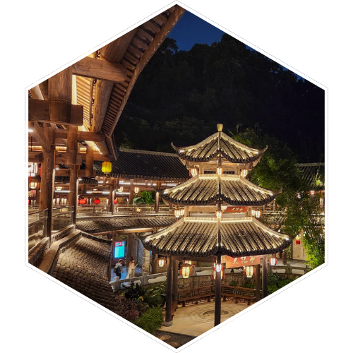 Night view of a traditional Chinese pavilion with multi-tiered, curved roofs illuminated by lights, surrounded by wooden walkways and buildings, with trees and a dark sky in the background.