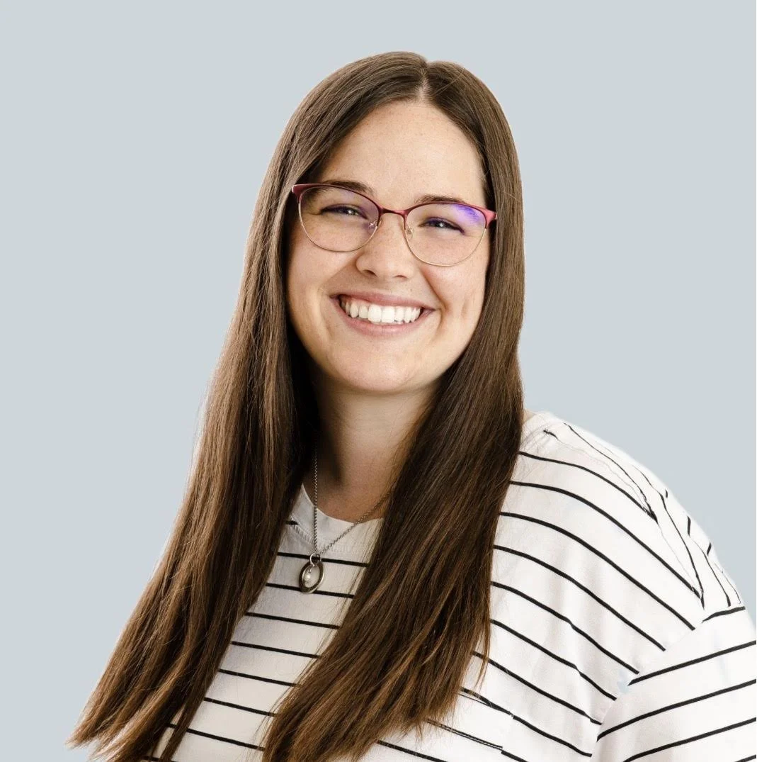 A young woman with long brown hair and glasses, smiling, wearing a white and black striped shirt and a necklace.