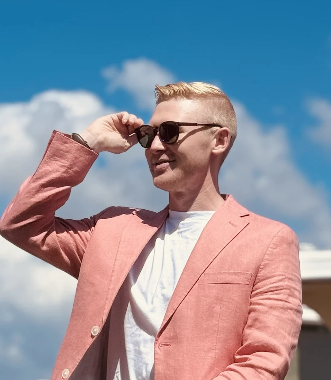 Chase Herd with blonde hair wearing sunglasses, a pink blazer, and a white shirt outdoors on a sunny day with a blue sky and white clouds.