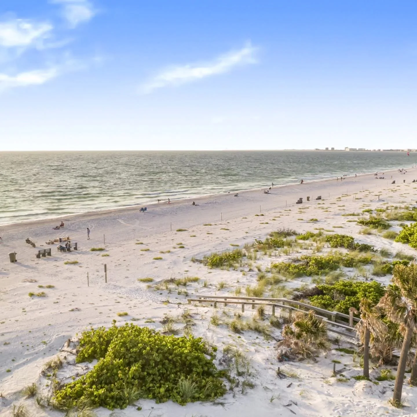 Beach with white sand, green vegetation, and a few people walking or sitting near the shoreline, with a calm ocean and blue sky in the background. St. Pete Beach