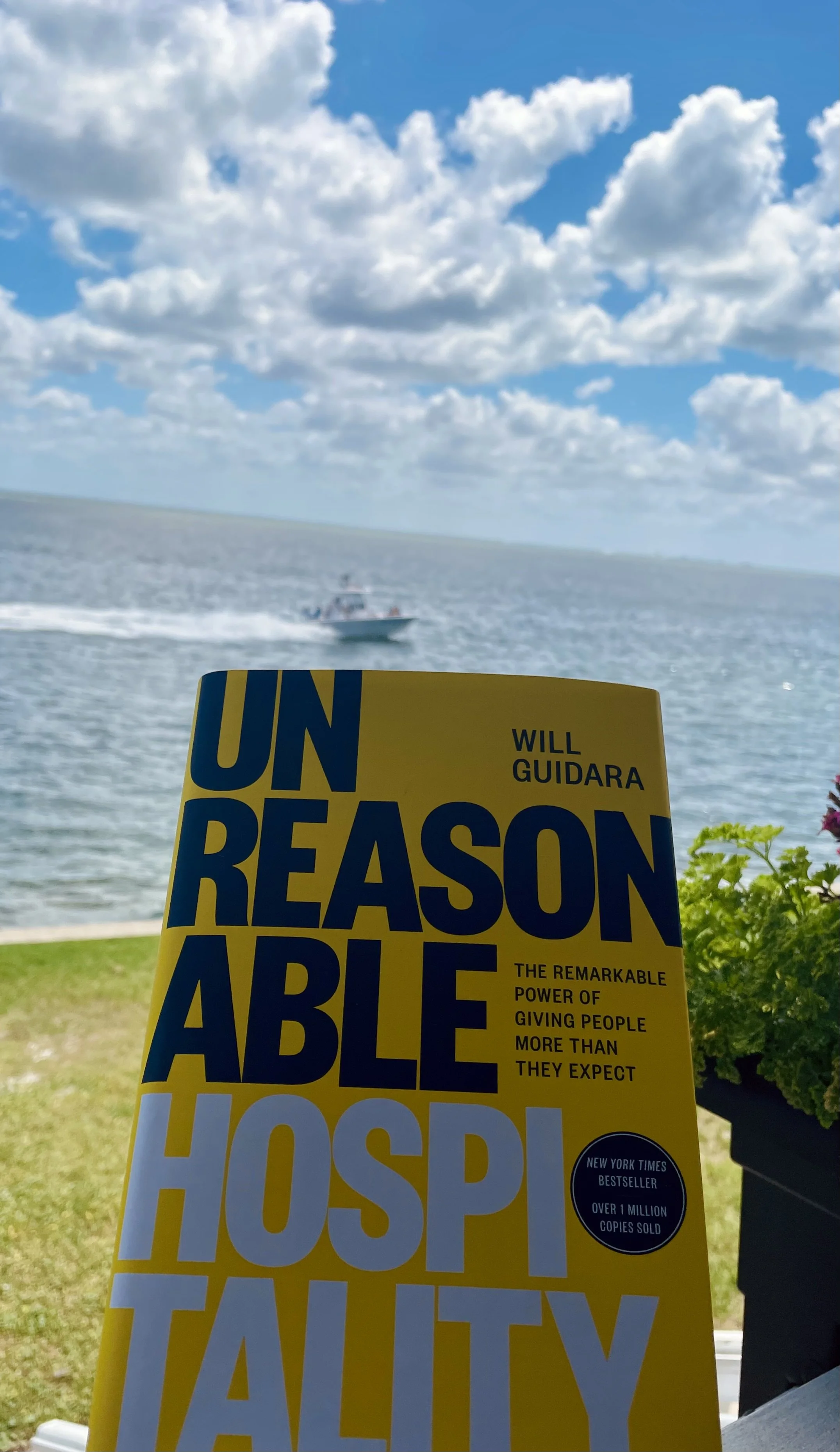 Cover of the book "Unreasonable Hospitality" by Will Guidara with a lakeside view and a boat in the water under a cloudy sky.