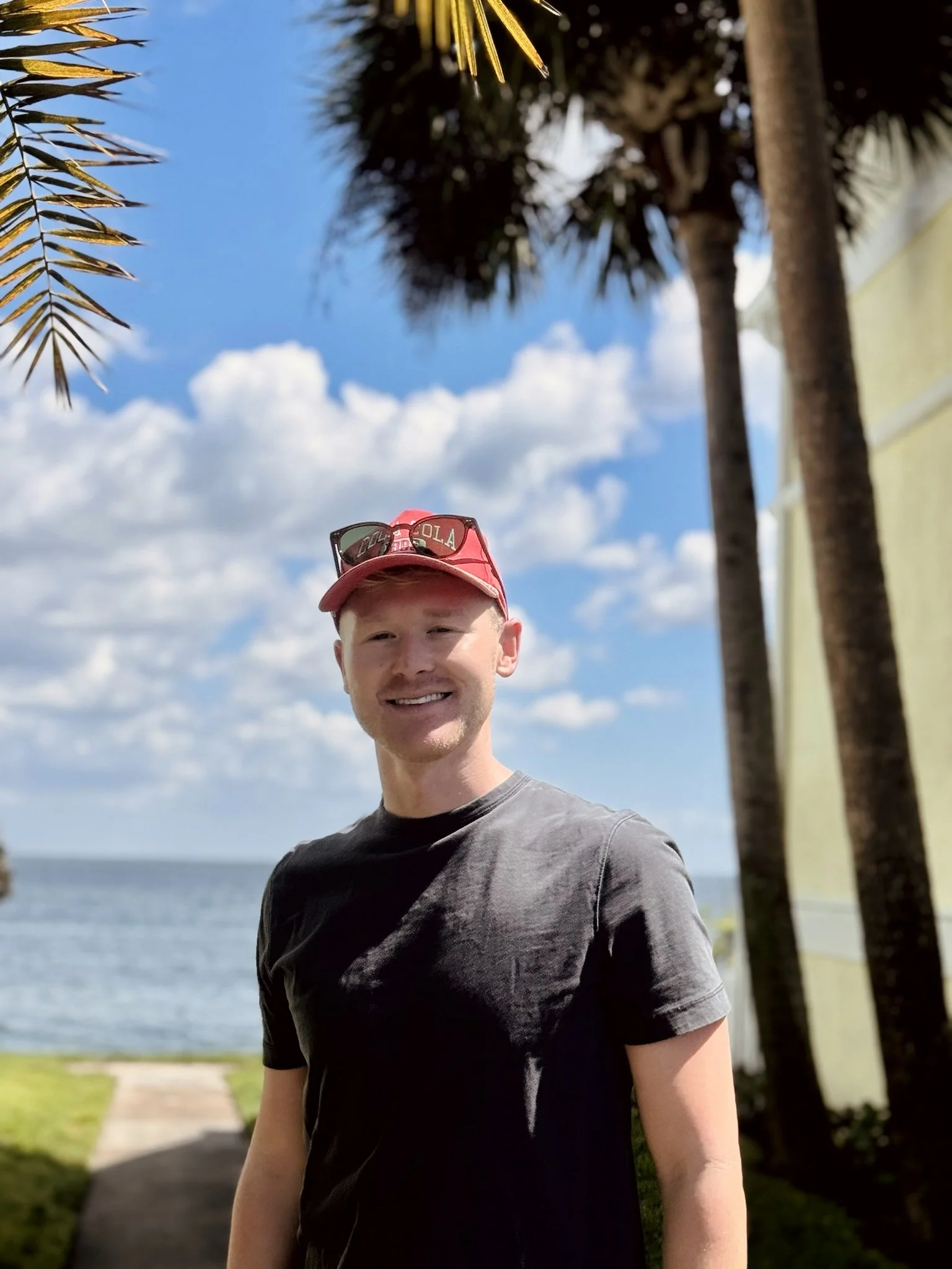 A young man smiling outdoors near the beach, with palm trees, a blue sky with clouds, and the ocean in the background. Chase Herd St. Petersburg, FL REALTOR