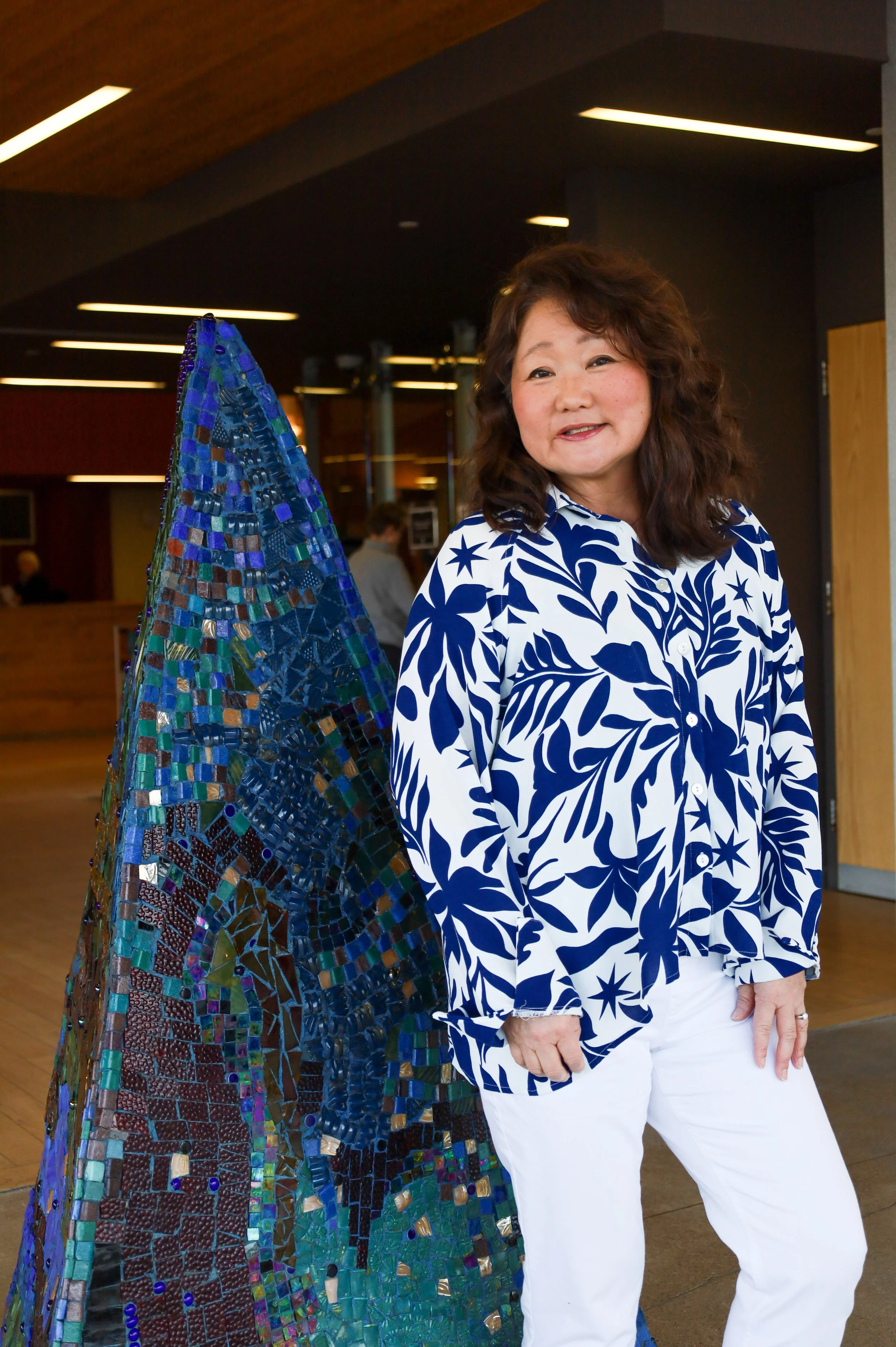 Sandy wearing a white and blue patterned blouse and white pants, standing next to a large mosaic art piece at the Traverwood Library.
