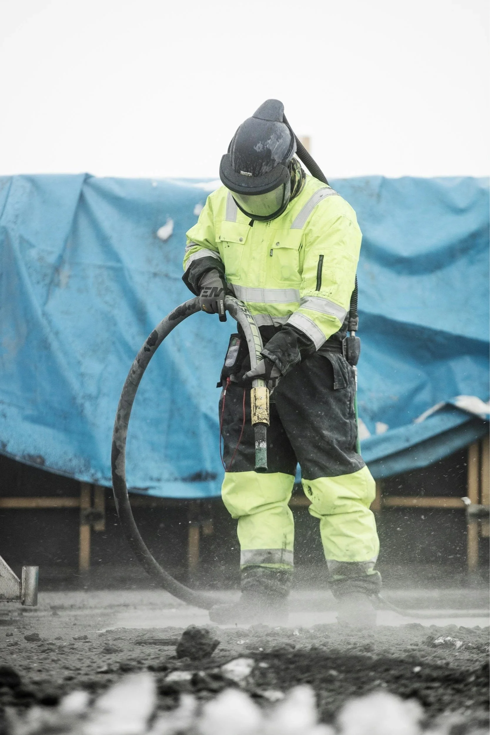 Worker in high-visibility yellow safety jacket and pants, wearing a black helmet, working with a large hose on a construction site, with a blue tarp in the background and dirt on the ground.