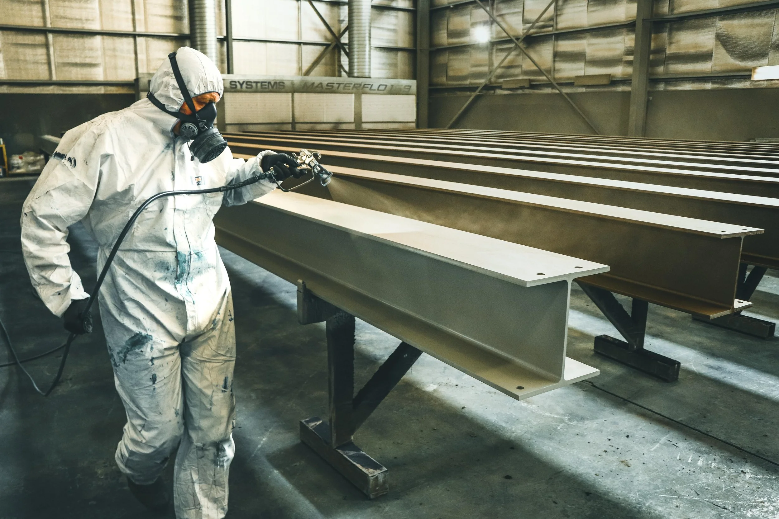 A worker dressed in protective white clothing, gloves, and a respirator mask is spray painting or coating large metal beams inside an industrial facility.