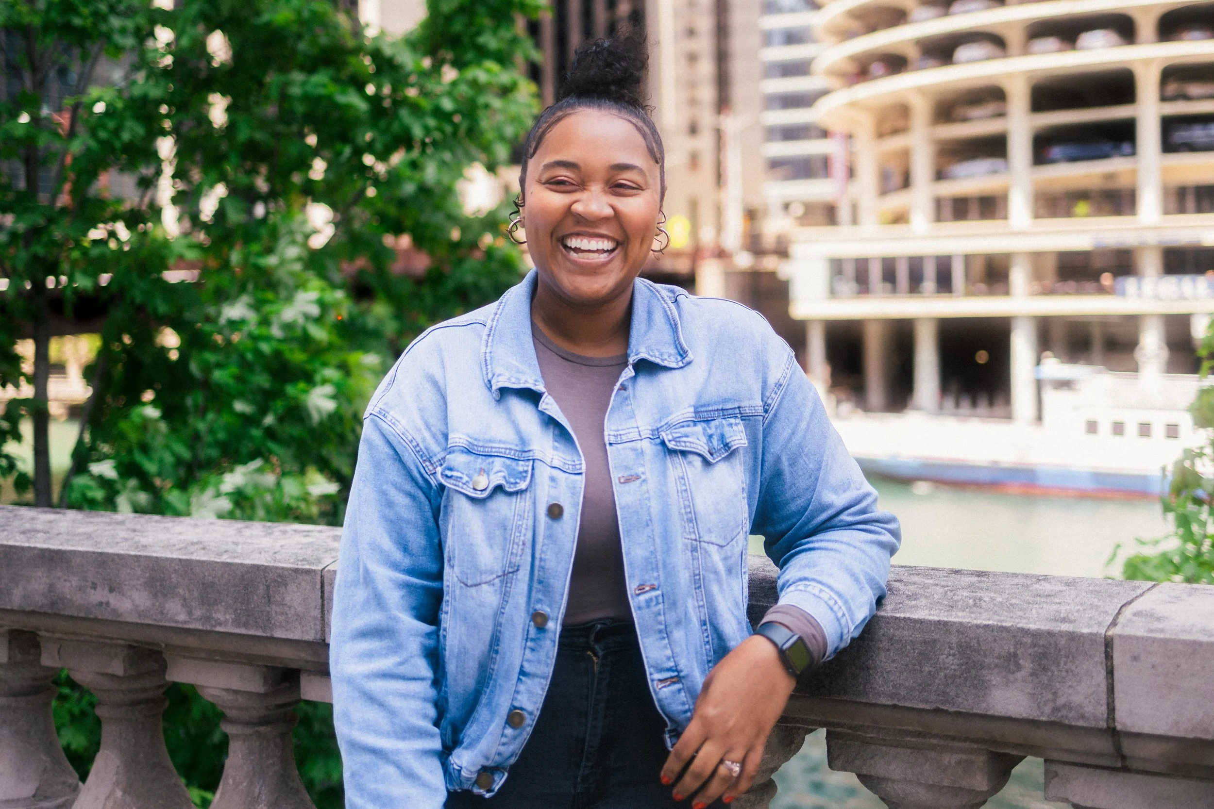 A woman with braided hair and hoop earrings is smiling and wearing a denim jacket, standing outdoors by a stone railing with a cityscape and water in the background.