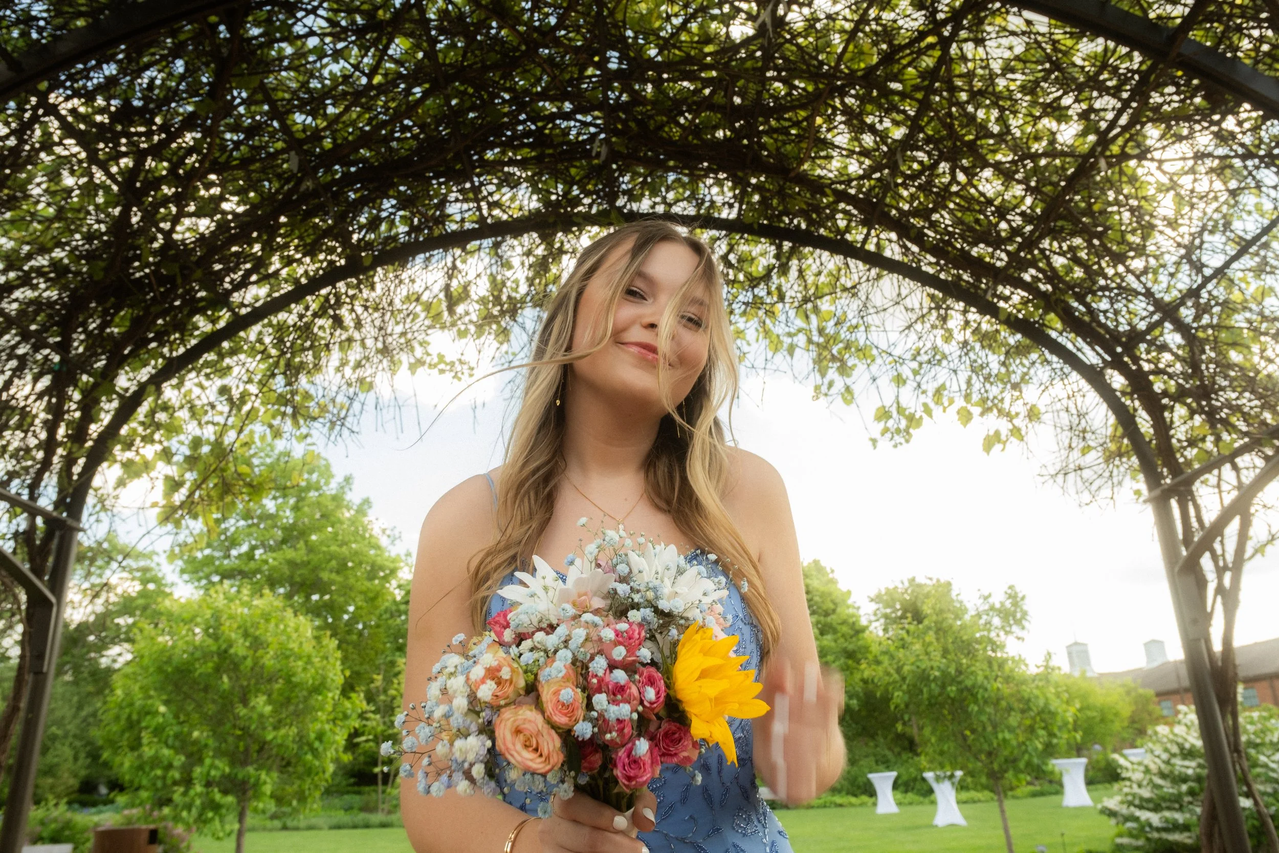 A young woman holding a bouquet of flowers, standing outdoors under a decorative arch made of intertwined branches and leaves, with trees and grassy area in the background.
