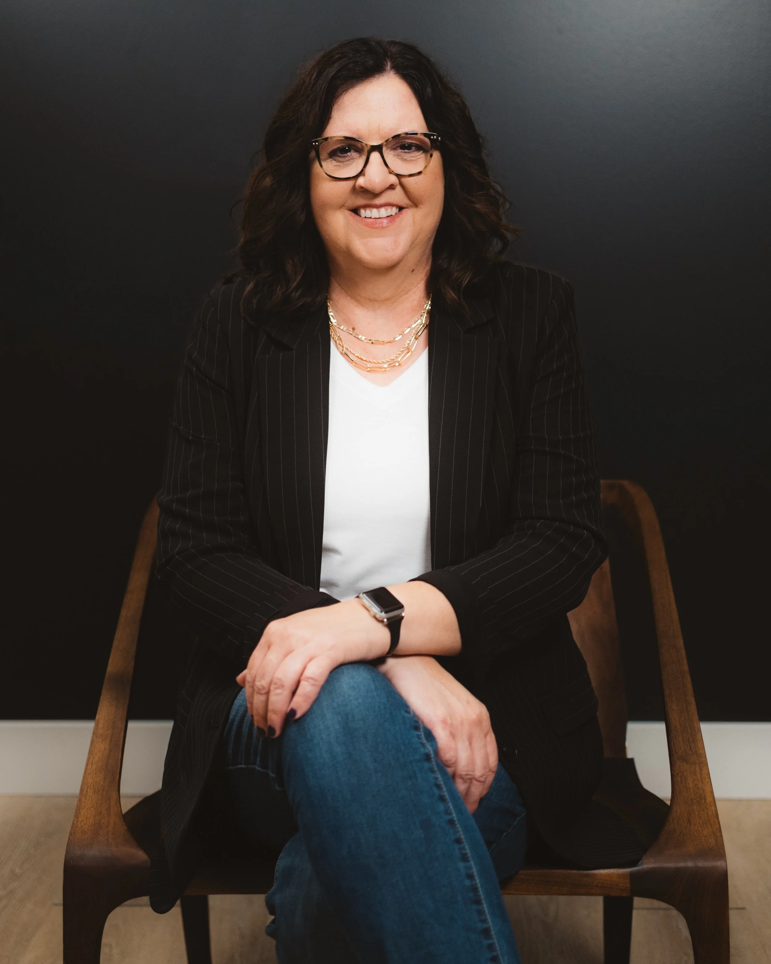 A woman with curly dark hair, glasses, and a friendly smile sitting on a wooden chair against a black background. She is wearing a black pinstripe blazer, a white shirt, jeans, jewelry, and a smartwatch.