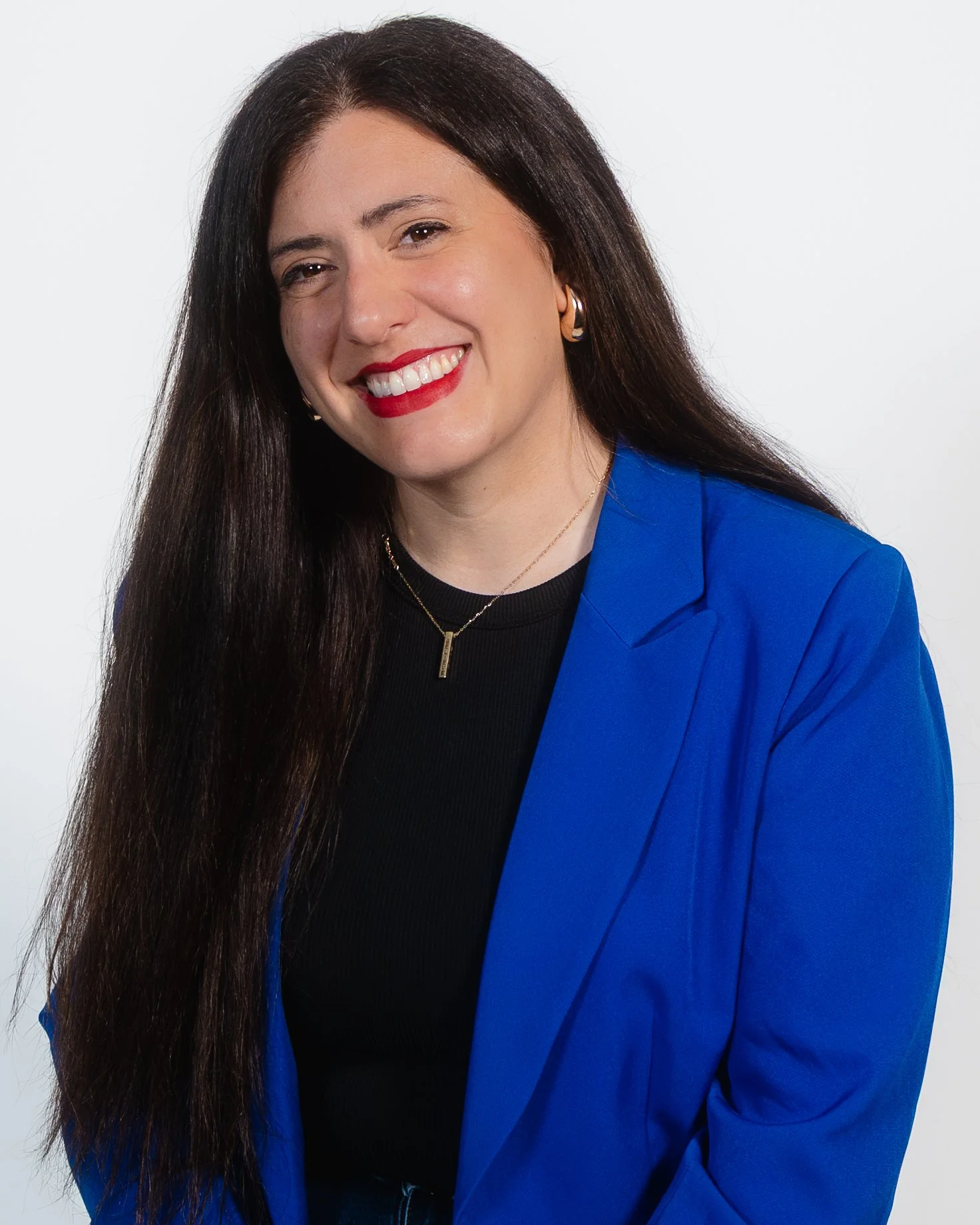 A woman with long dark hair, smiling, wearing a blue blazer, black top, gold jewelry, and red lipstick, against a plain white background.