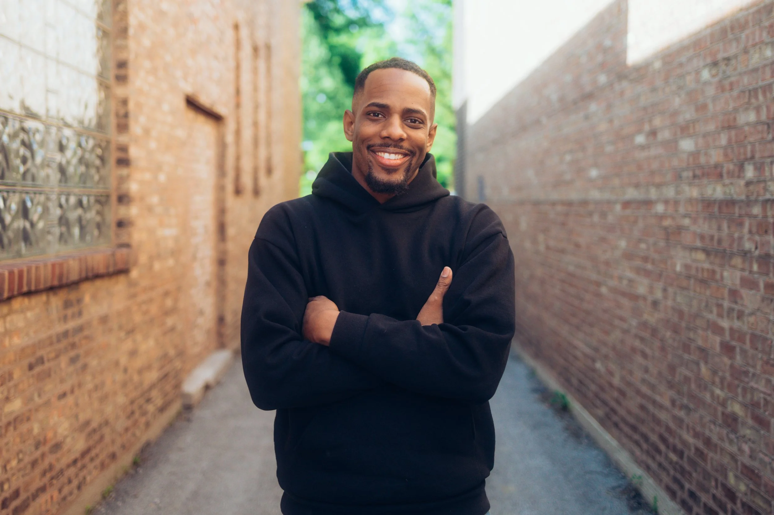 A young man with a beard smiling, standing with crossed arms in an alleyway between brick buildings, wearing a black hoodie.