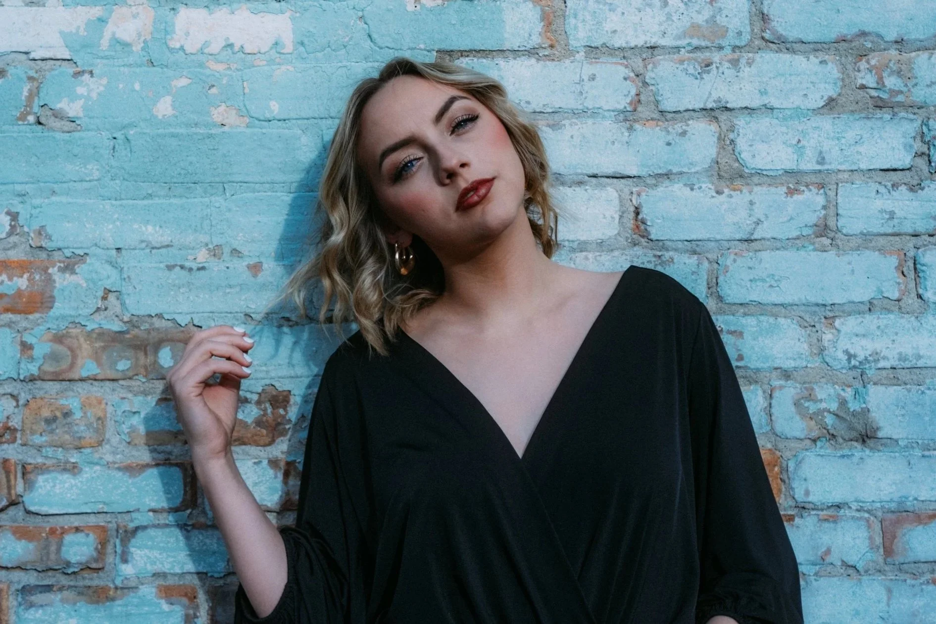 A woman with blonde, wavy hair and hoop earrings posing against a blue and red brick wall, wearing a black V-neck top.