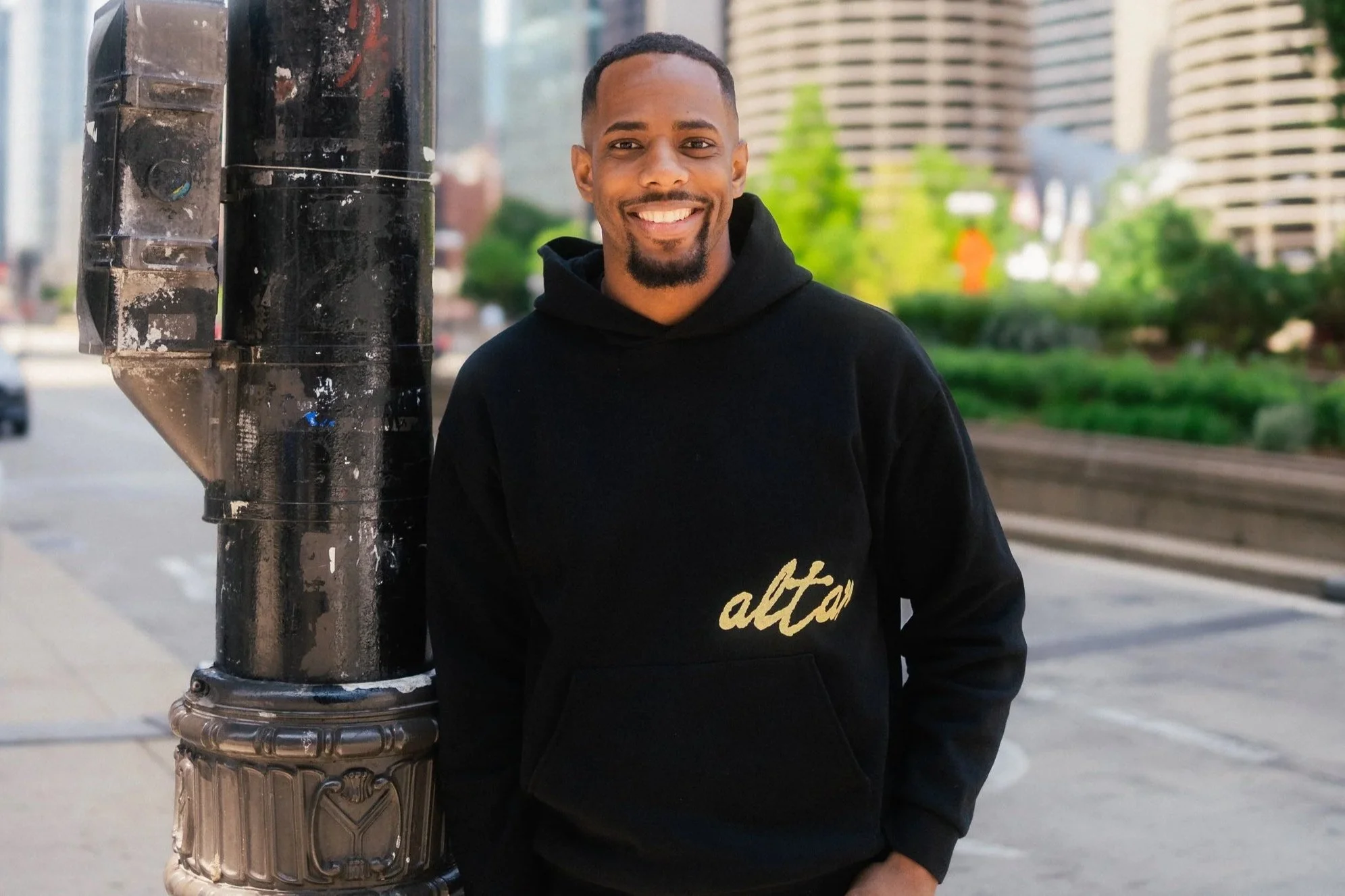 A smiling man wearing a black hoodie with the word 'alt' in gold lettering, standing next to a black lamppost on a city sidewalk with green trees and high-rise buildings in the background.