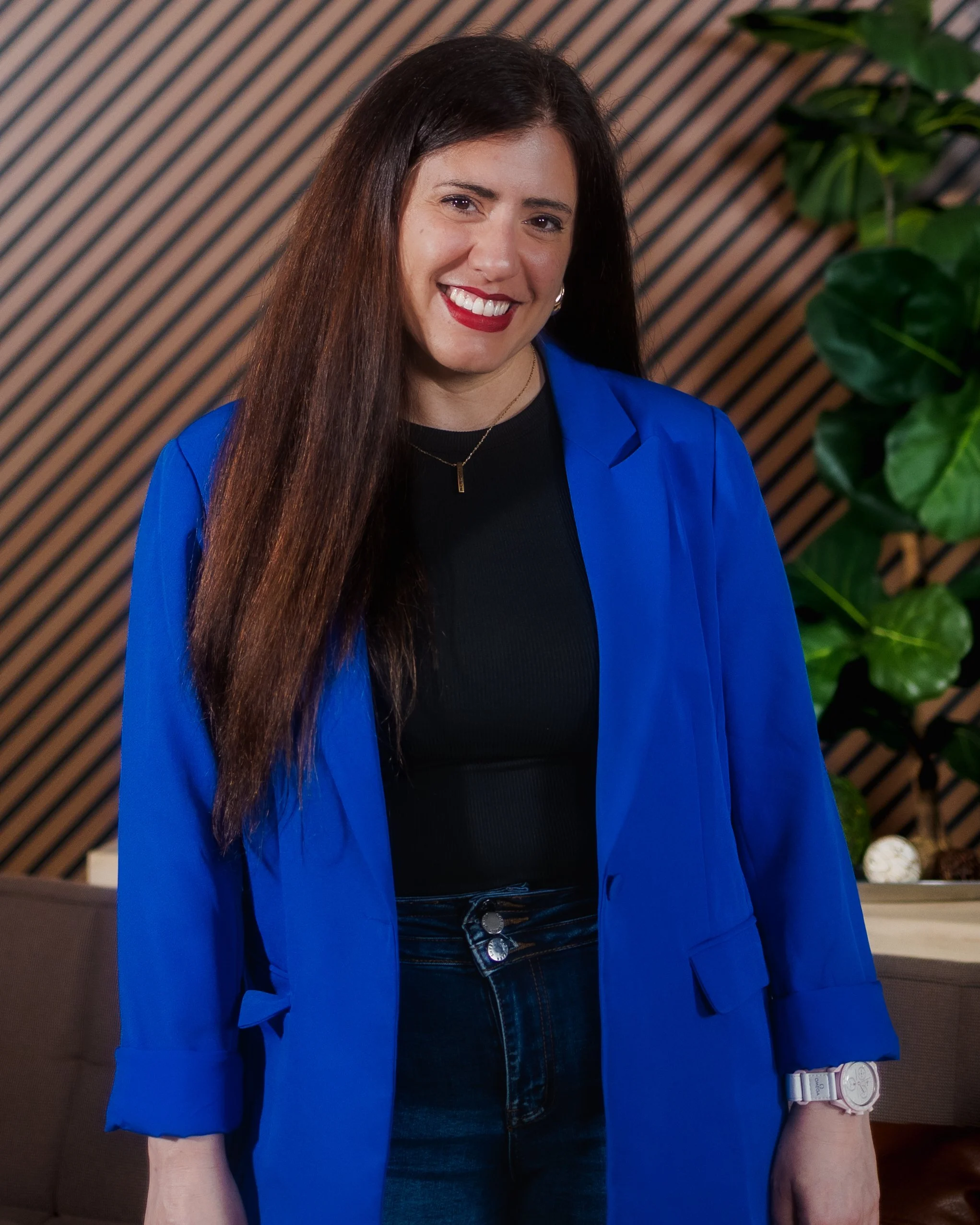 A woman with long brown hair wearing a bright blue blazer, black top, and dark jeans, smiling and standing indoors with a wooden panel backdrop and green leafy plant.