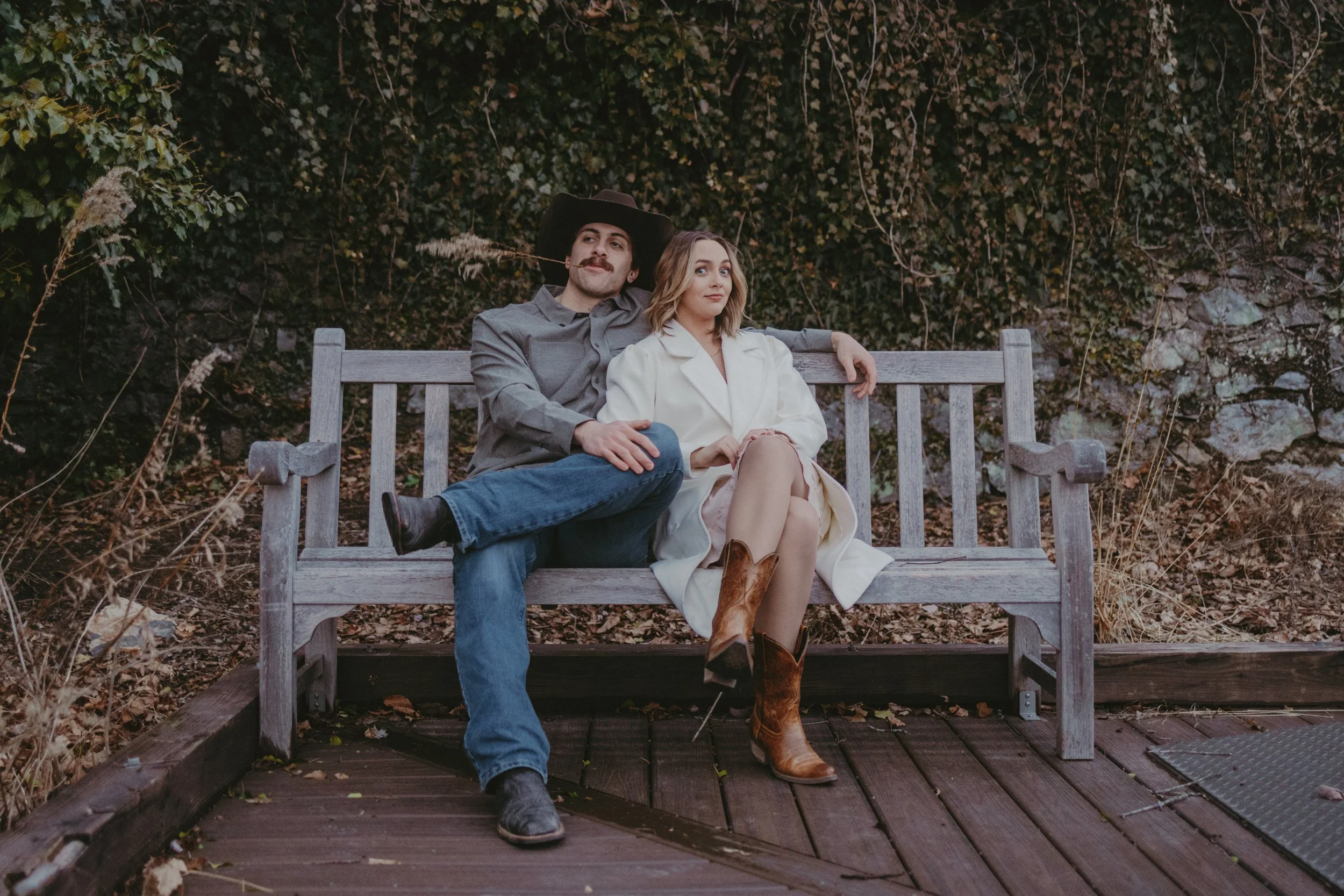 A man and woman sitting on a wooden park bench in front of bushes and rocks. The man wears a cowboy hat, gray shirt, and jeans, with his arm around the woman. The woman wears a white coat and cowboy boots, sitting with her legs crossed.