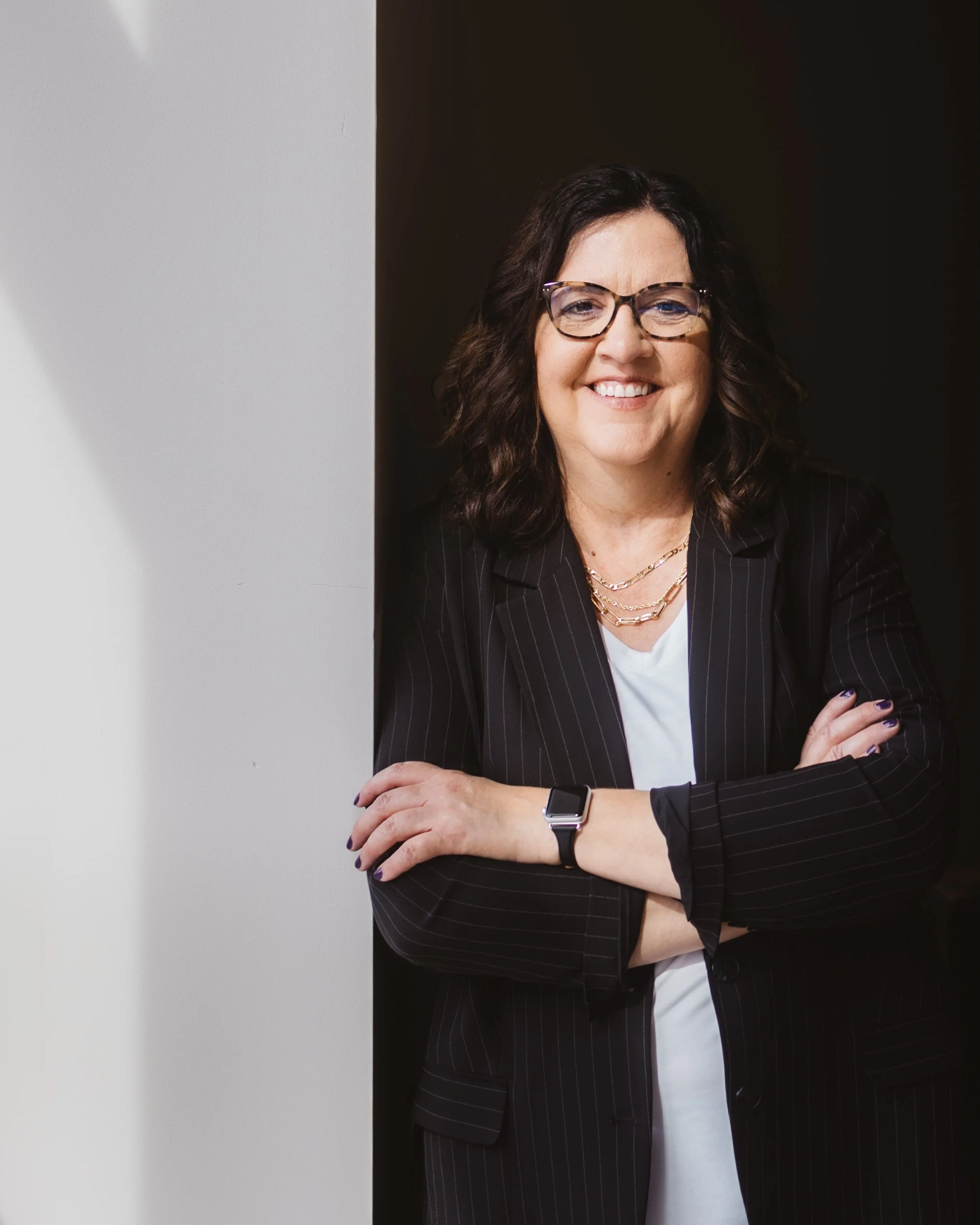 Woman with glasses smiling, wearing a black pinstripe blazer and white shirt, standing with arms crossed, against a white and dark background.