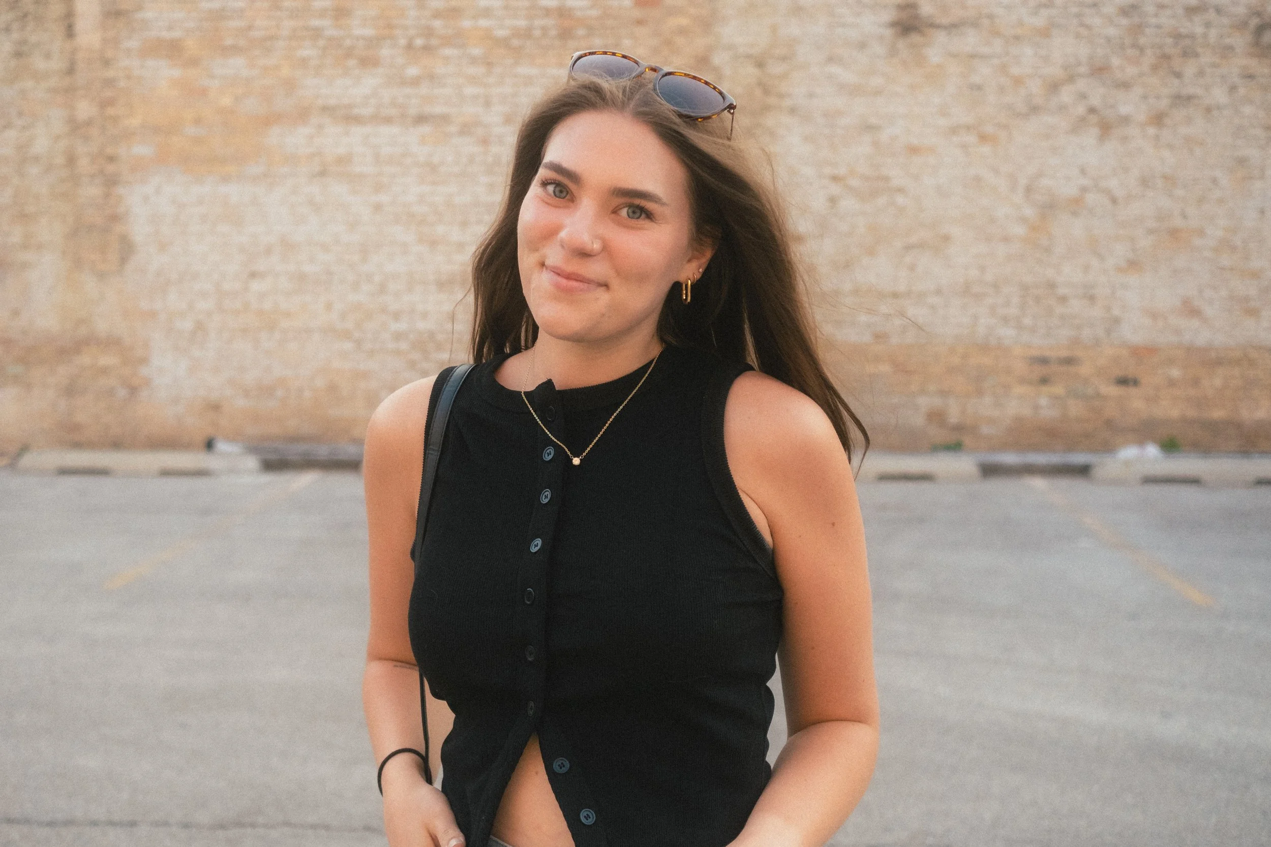 A young woman with long brown hair, wearing a sleeveless black top, sunglasses on her head, gold hoop earrings, a necklace, and a black wristband, stands outdoors in front of a brick wall and parking lot.