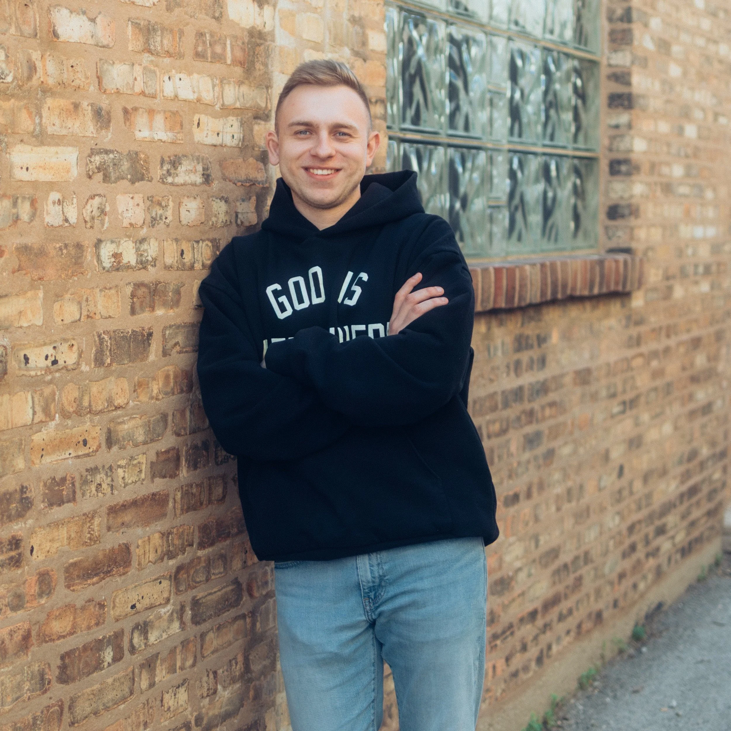 A young man with short light brown hair, smiling, standing against a brick wall outside. He is wearing a black hoodie with white lettering and light blue jeans.
