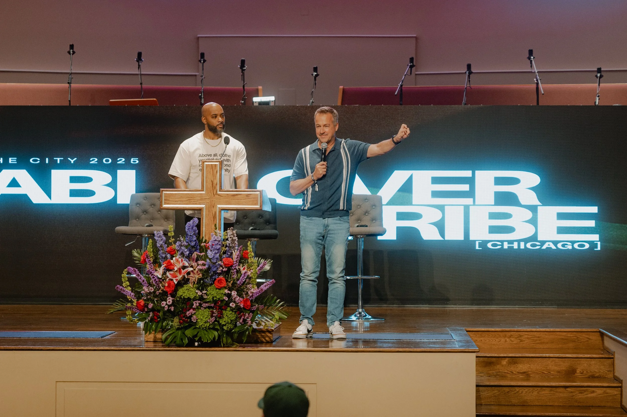 Two men on stage at a church event, one speaking into a microphone and the other looking on, with a large cross and flower arrangement in front of them and a big screen behind displaying 'Summer Tribe' and 'Chicago'.