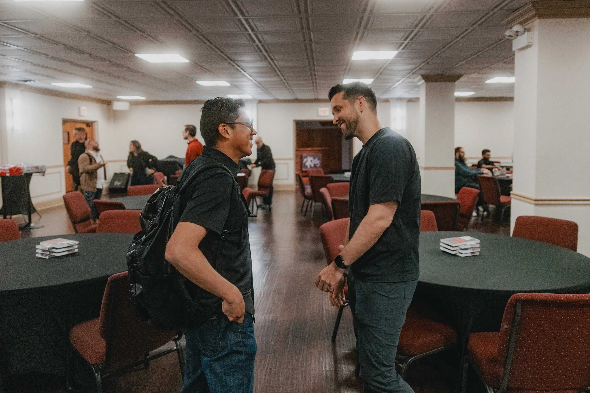 Two men smiling and facing each other in a conference room, engaging in a friendly conversation. The room has round tables covered with black cloths and chairs, with some people in the background.