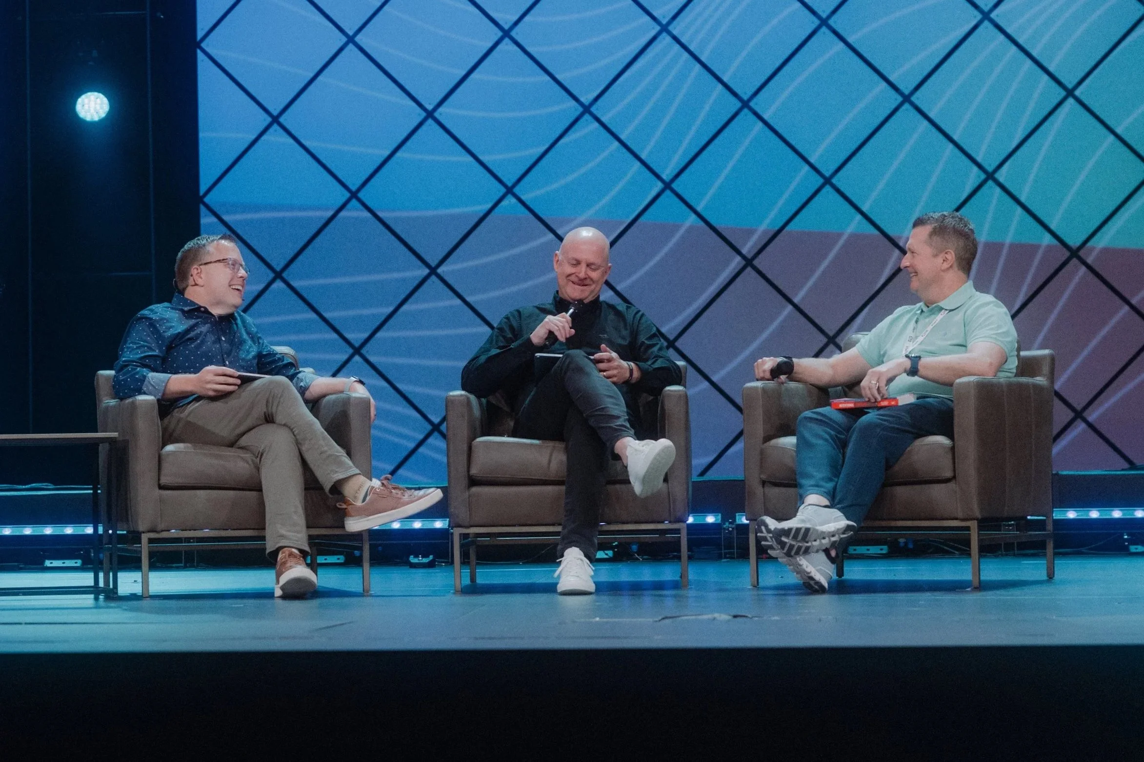 Three men sitting in chairs on a stage, engaged in conversation and smiling, behind a blue patterned backdrop.