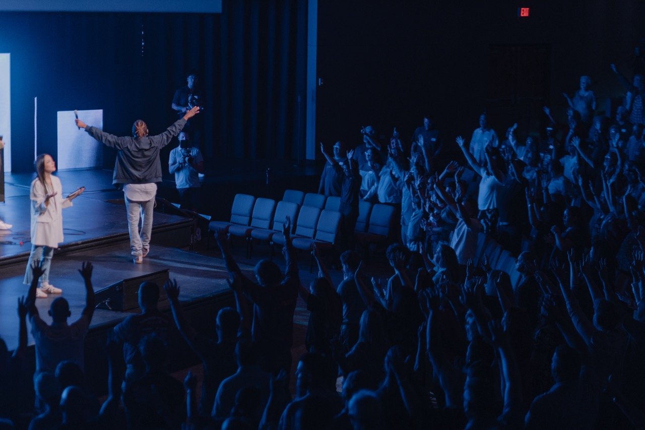 People on stage leading a worship or motivational event with their hands raised, audience members in front with hands up, in a darkened auditorium with blue lighting.