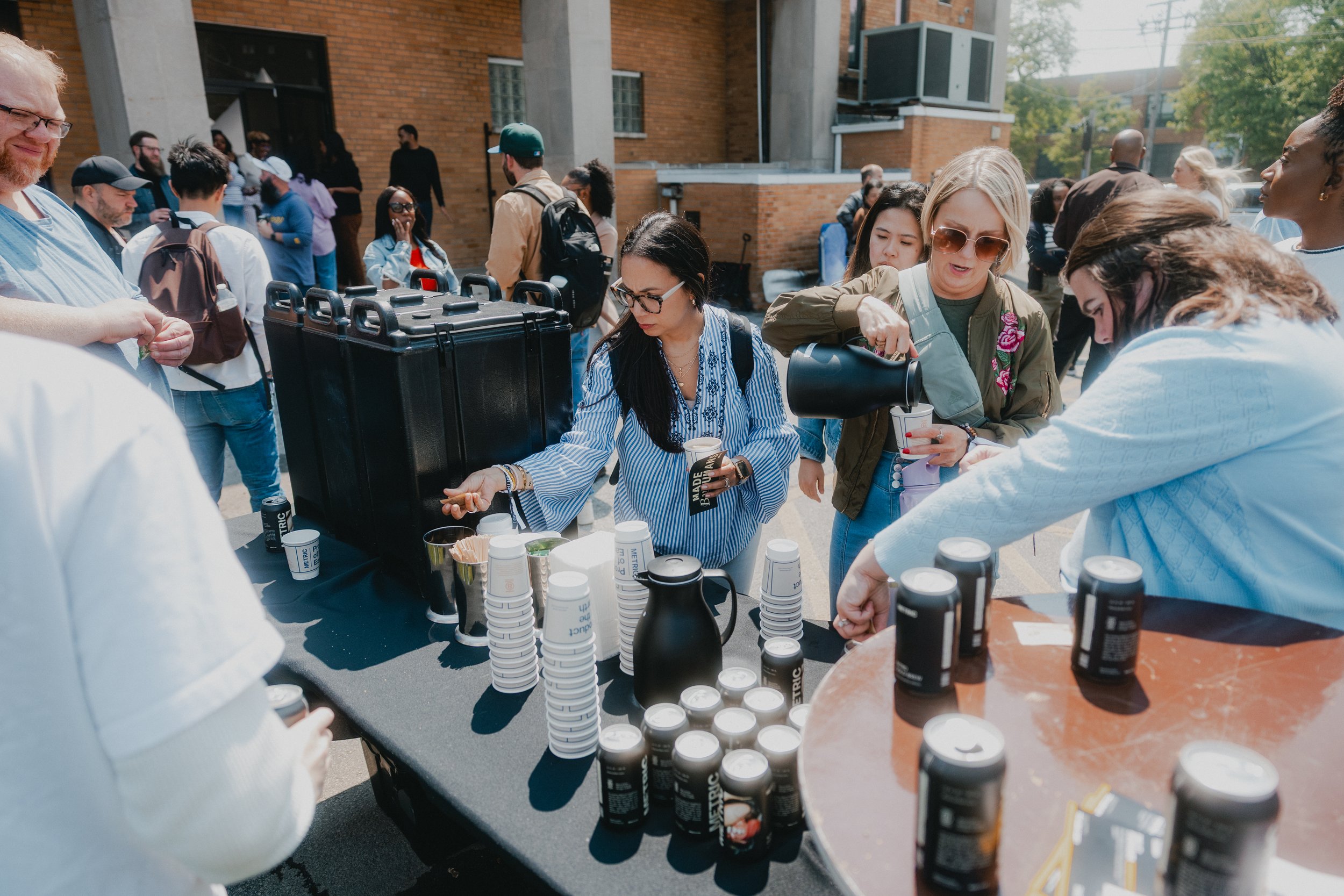 People gathering at an outdoor event, getting drinks from a table with coffee cups, cans, and a coffee dispenser, on a sunny day in front of a brick building.