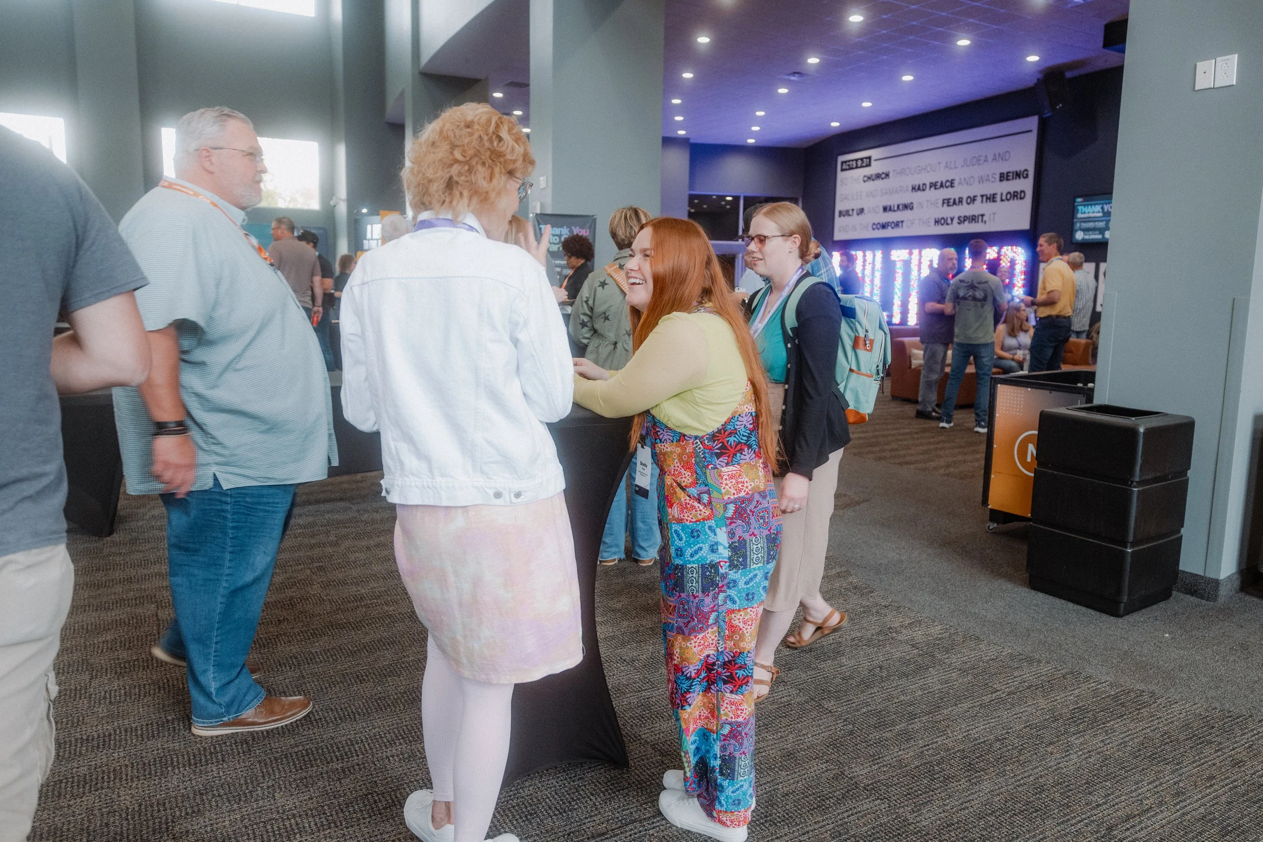 People socializing at a conference or event, standing around a high table in a crowded, modern indoor venue.