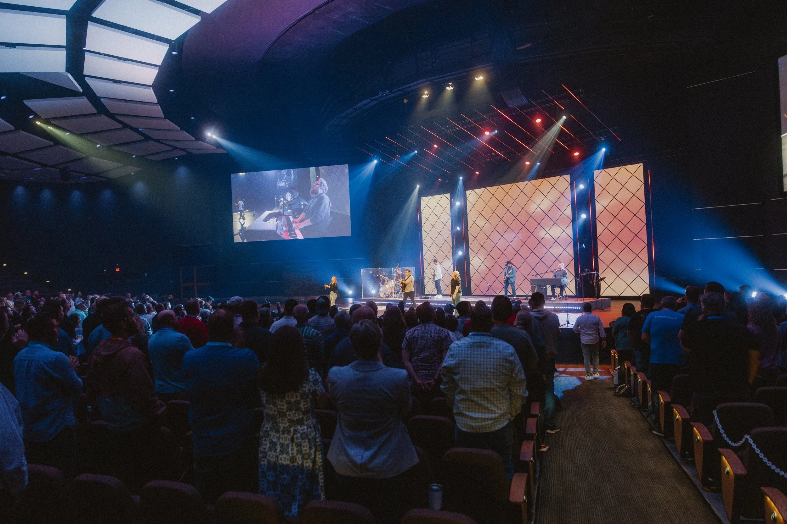 Crowd attending a live concert or church service with a large stage, multiple performers, and colorful lighting effects.