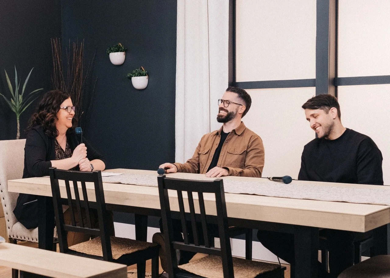 Three people in a panel discussion at a table, two men and a woman, all smiling and holding microphones, in a modern room with plants on the wall.