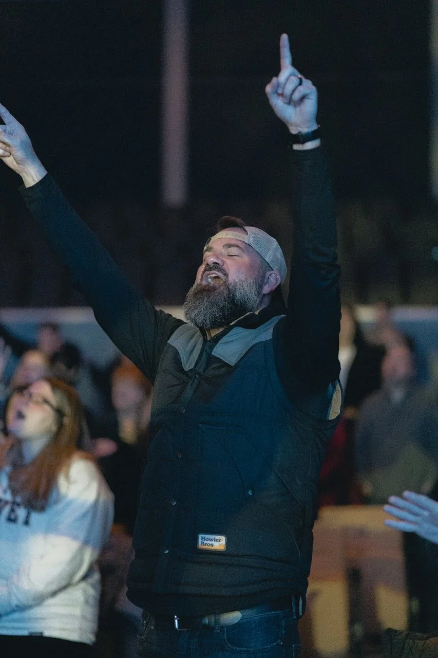 A bearded man wearing a cap backwards and a black vest raising his arms and eyes closed at an outdoor gathering or concert.