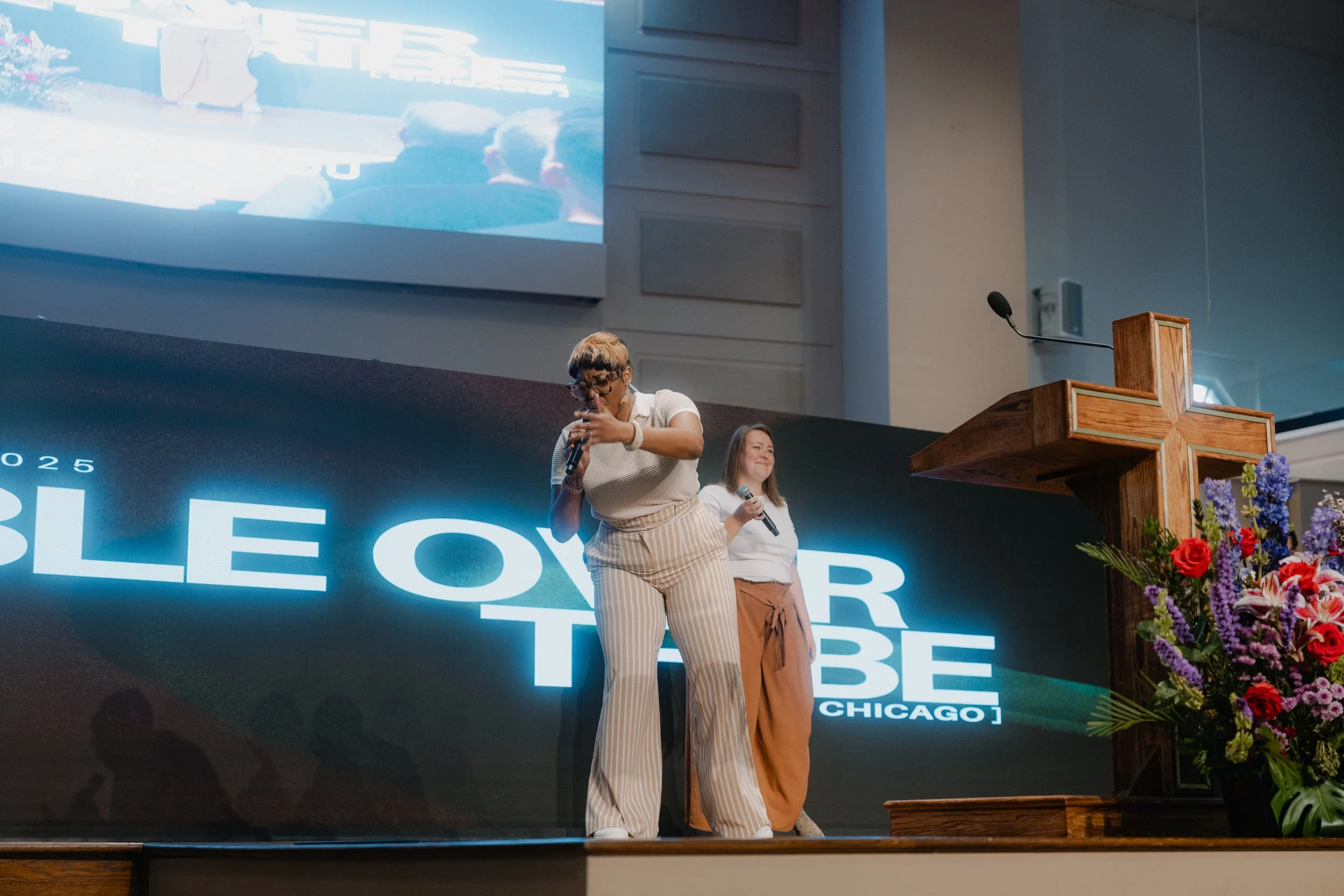 Two women standing on stage, one holding a microphone and pointing down, the other smiling with a microphone in hand, behind a podium with flowers, in a church or conference hall with a large screen displaying the words "Bible Over Tube Chicago."