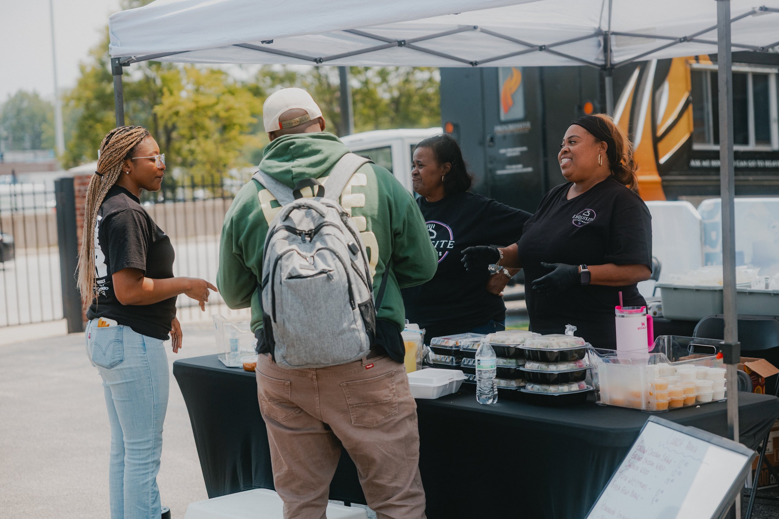 People standing at a food stall outdoors, possibly at a market or festival, with food containers and bottled water on the table.
