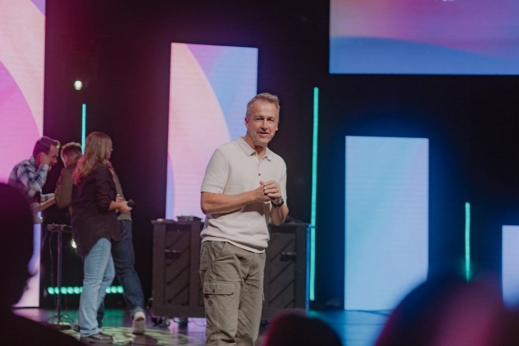 Man speaking on stage with a group of people behind him in a dark lit environment with colorful illuminated screens.