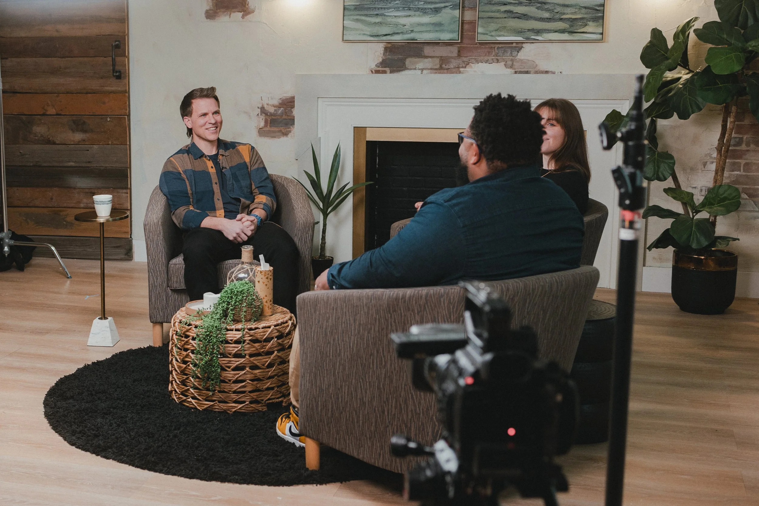 Three people engaged in an interview or podcast recording in a cozy living room setting. One person is speaking on a chair, while the other two are sitting on a couch listening.