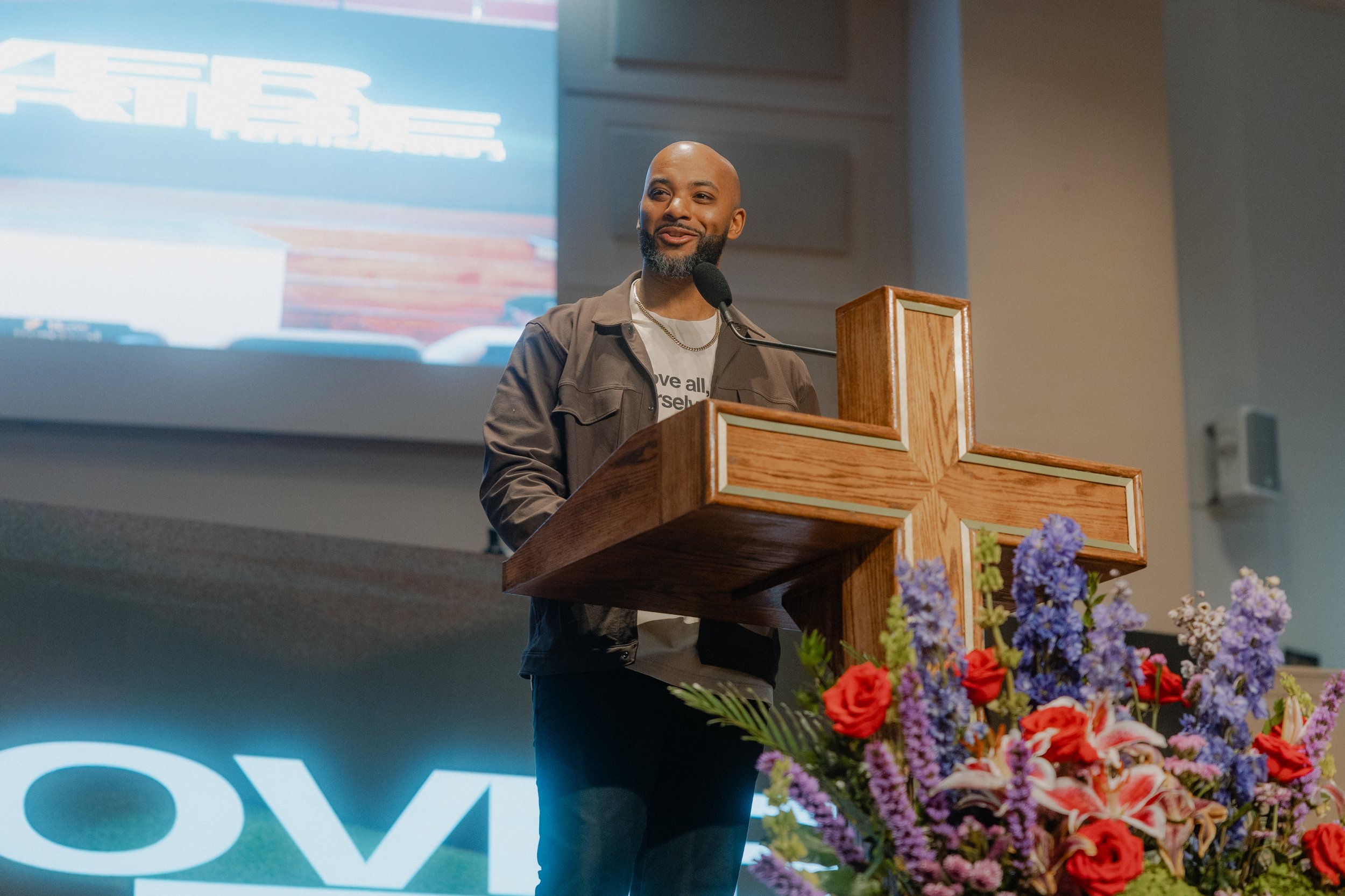 A man speaking at a church podium with a wooden cross, decorated with purple and red flowers in the foreground, and a large screen in the background.