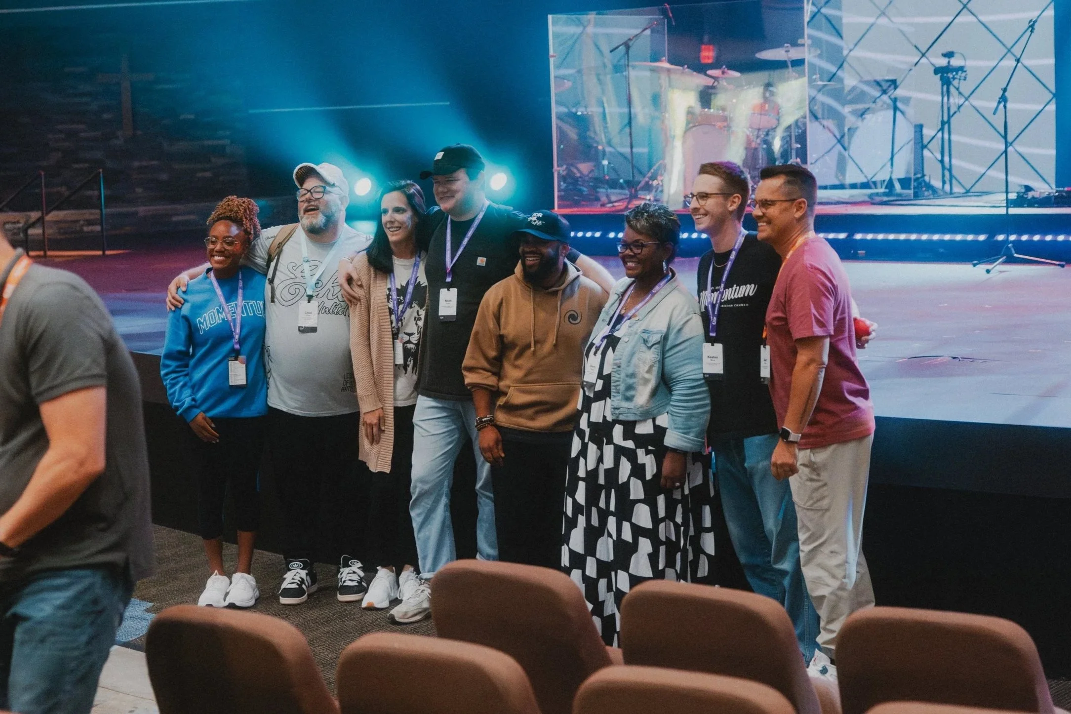 Group of nine people posing for a photo on stage at an event, with a drum set and musical instruments on a large screen behind them.