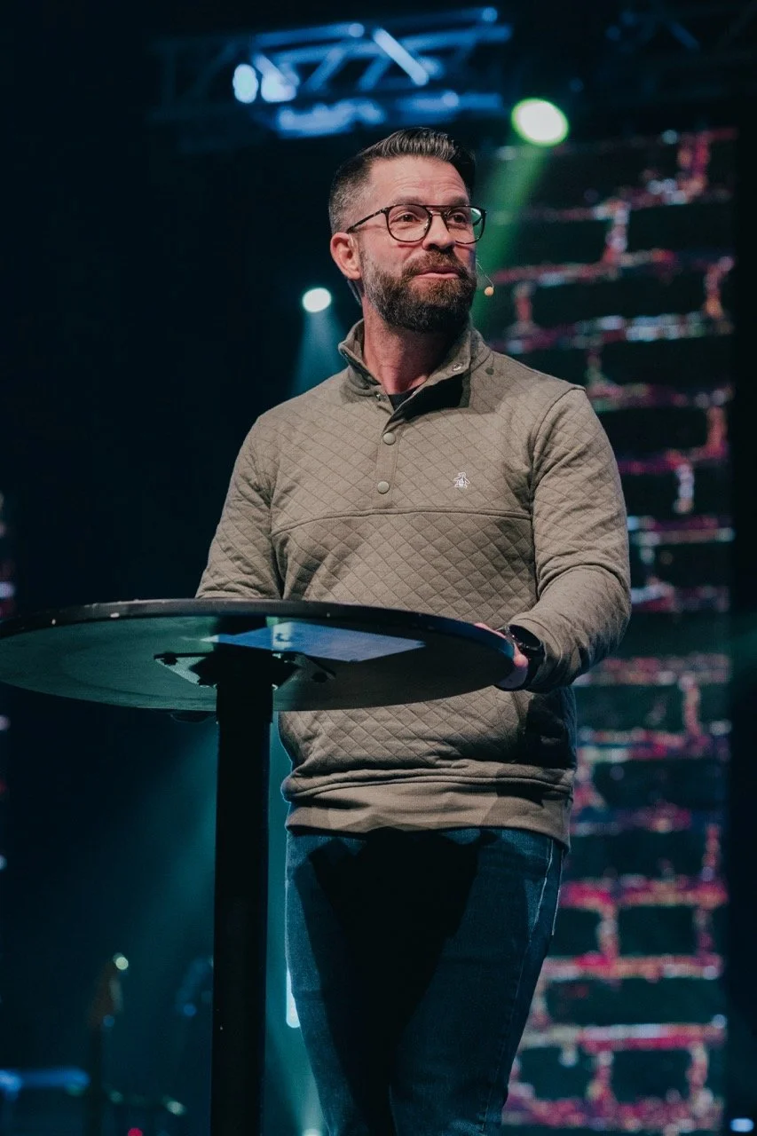 A man with glasses and a beard standing at a small round table on a stage with colorful lights and LED screens in the background.